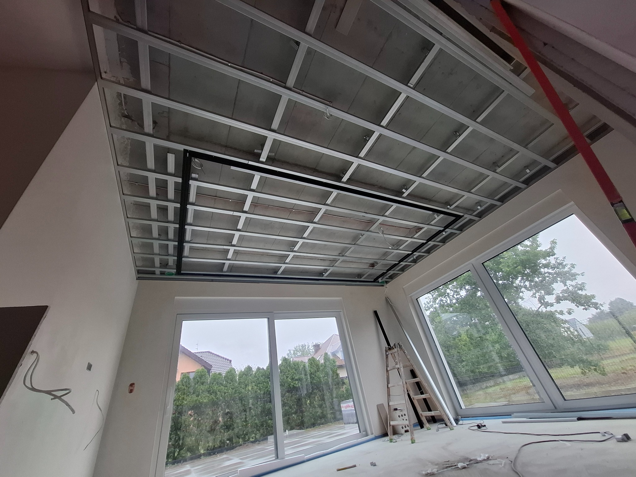 Sufit podwieszany z metalową konstrukcją w jasnym wnętrzu z dużymi oknami, drabina oparta o ścianę, widoczne przewody elektryczne.