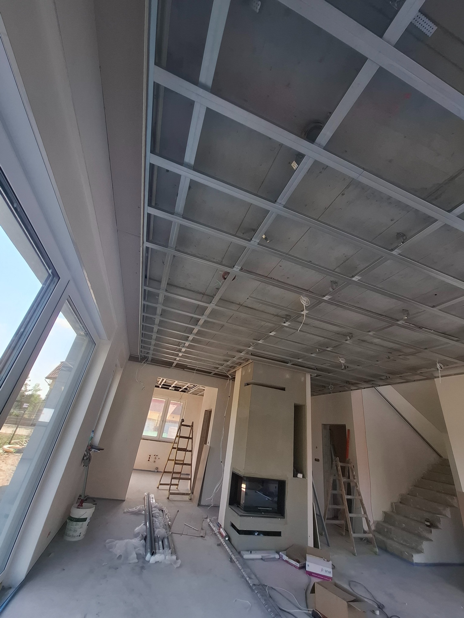 Montaż stelaża sufitu podwieszanego z profili w nowym domu w Warszawie. Widoczne elementy instalacji i kominek. W tle drabina i okno.