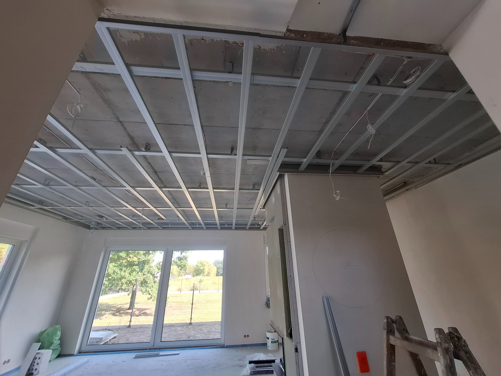 Widok na sufit z zamontowaną metalową konstrukcją pod płyty gipsowo-kartonowe w jasnym wnętrzu. Widoczne okna z widokiem na zieleń i elementy wyposażenia.