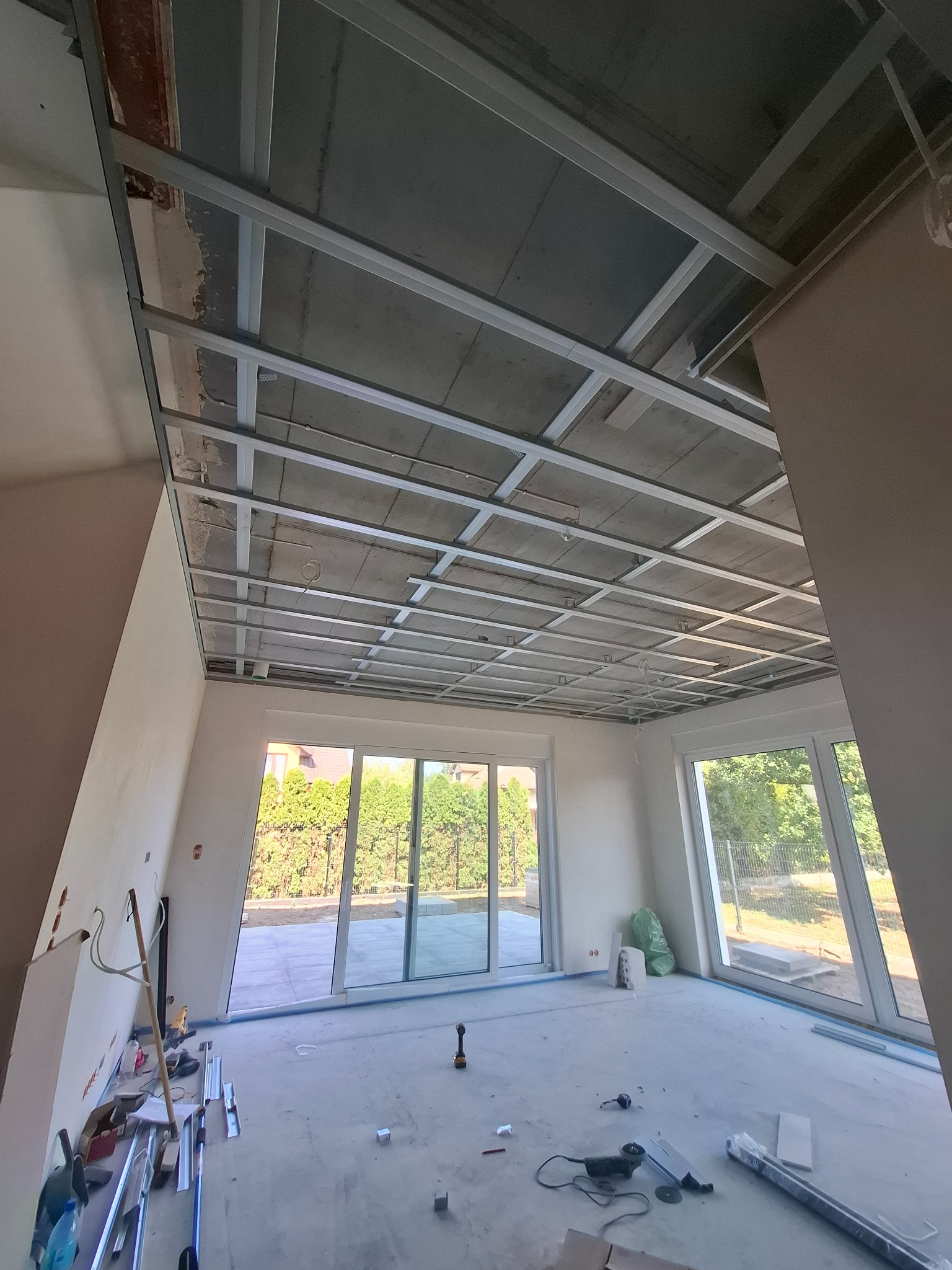 Montaż sufitu podwieszanego z metalowej konstrukcji w jasnym pomieszczeniu z dużymi oknami. Widoczne narzędzia i materiały budowlane.