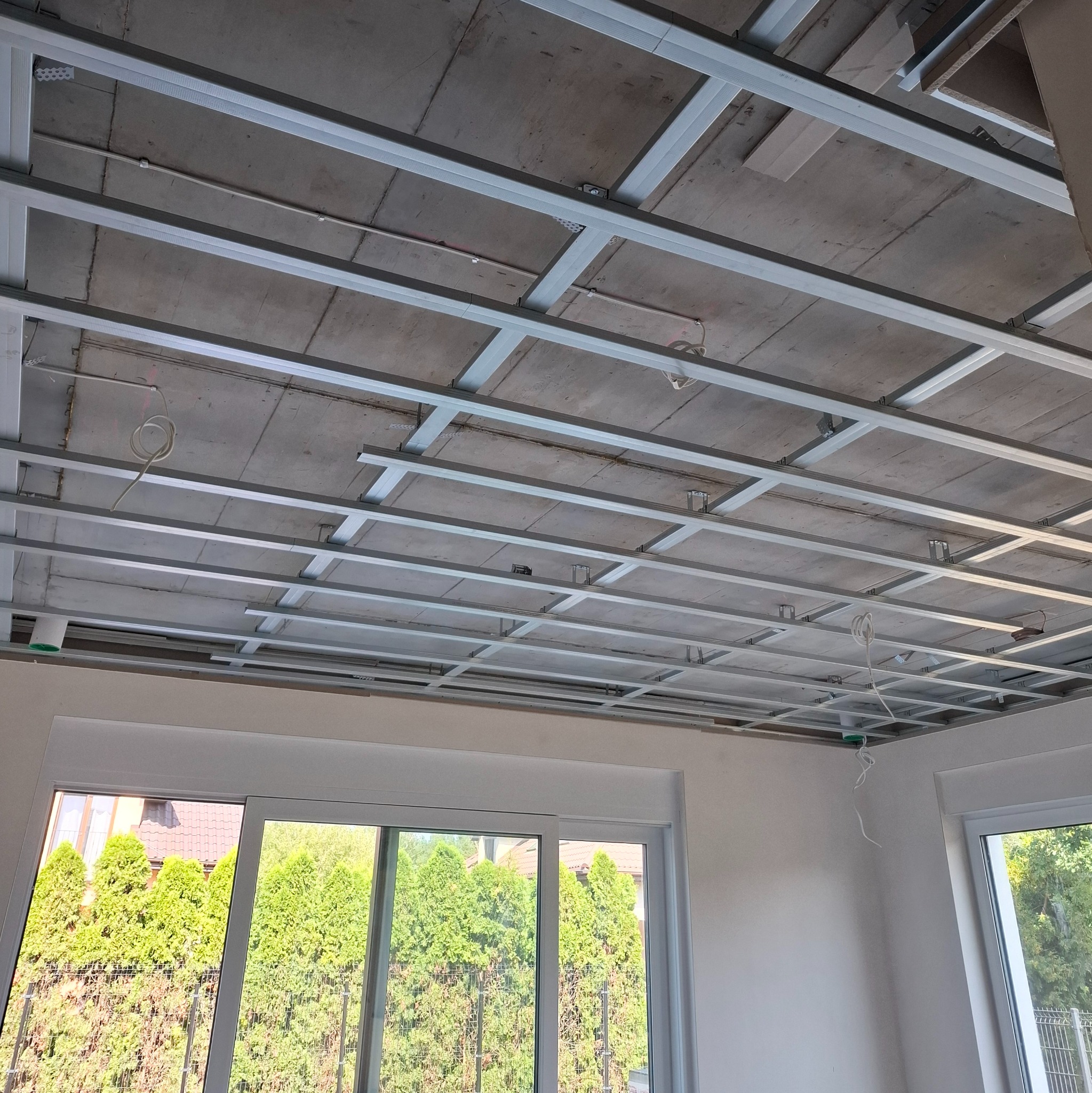 Montaż sufitu podwieszanego z płyt kartonowo-gipsowych, widoczne metalowe profile i częściowo zamontowane płyty. W tle okno z widokiem na zieleń.