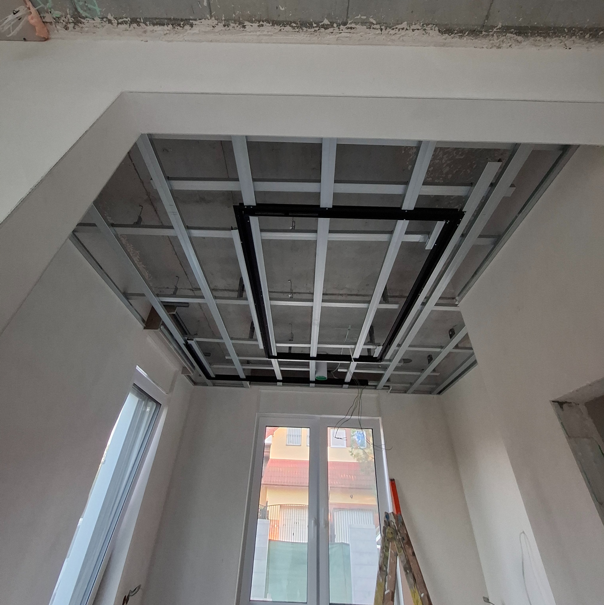 Sufit podwieszany z metalową konstrukcją i elementami instalacji, w trakcie montażu w jasnym pomieszczeniu z oknami i drabiną. Widok z dołu.