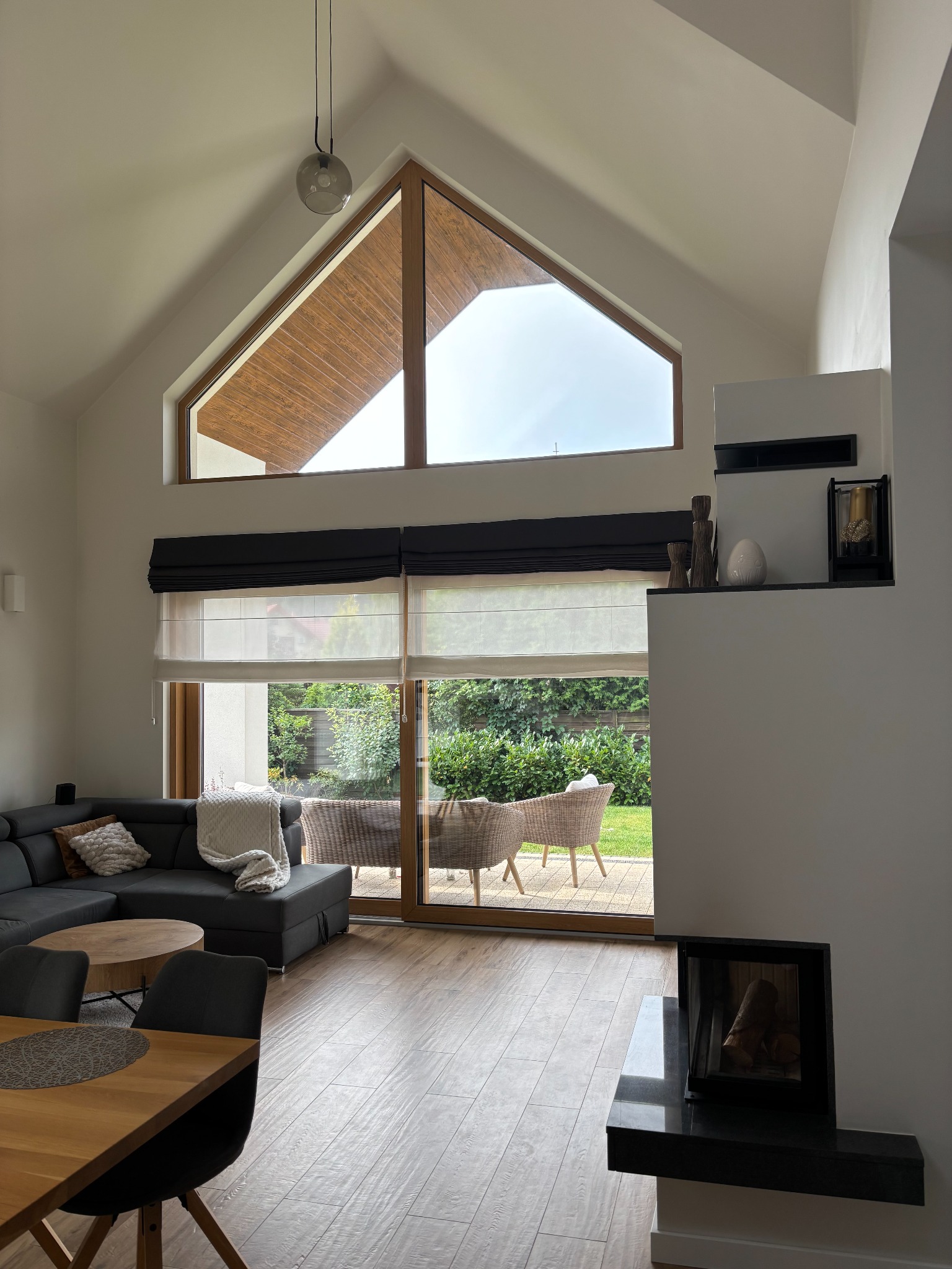 Salon z wysokim oknem i drewnianą podłogą. W tle widoczny ogród przez przesuwne drzwi. Minimalistyczny kominek i elementy dekoracyjne.