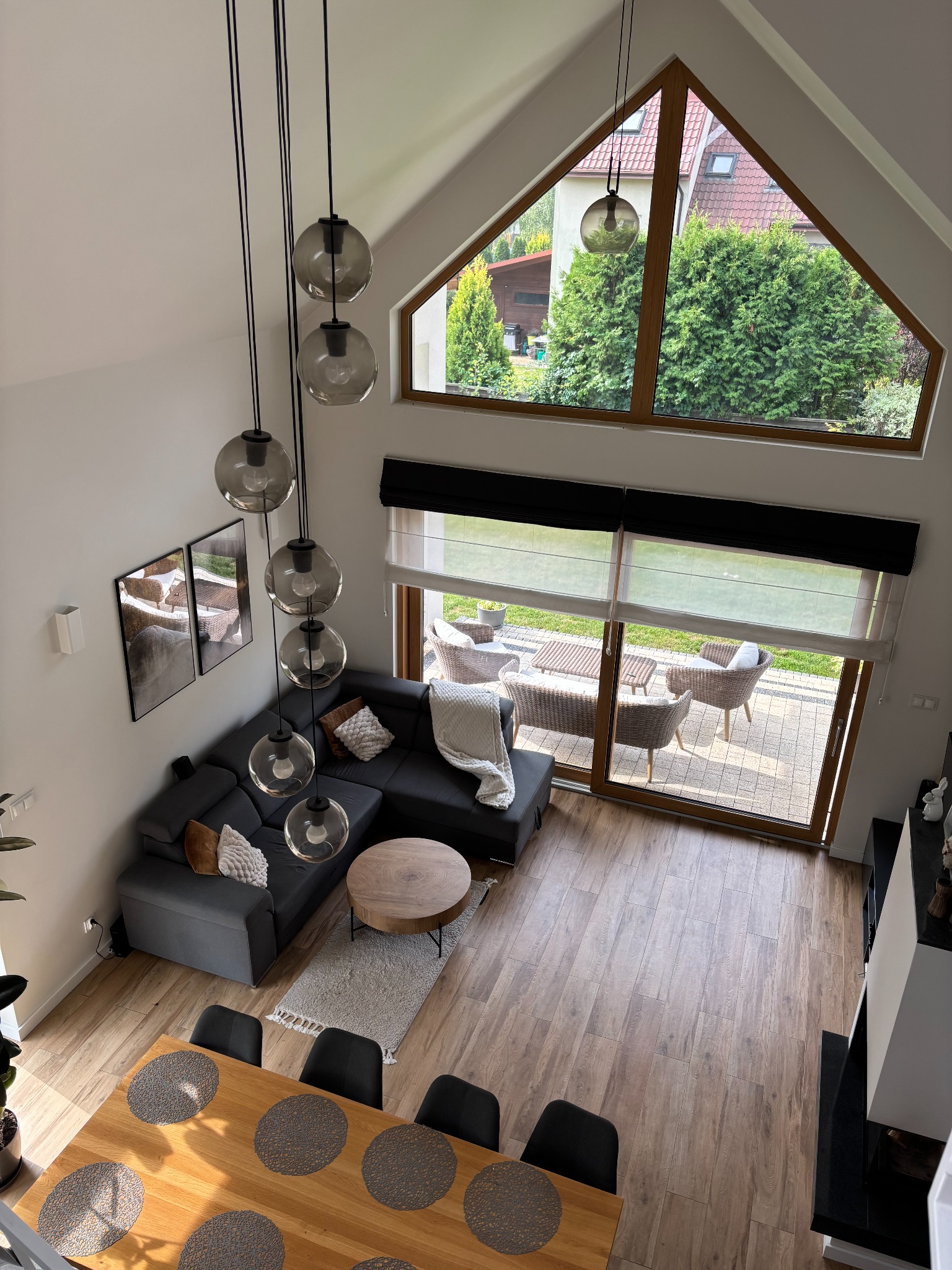 Wysoki salon z antresoli: szary narożnik, drewniany stół i lampy wiszące. Duże okno z widokiem na ogród. Parkiet na podłodze i jasne ściany.