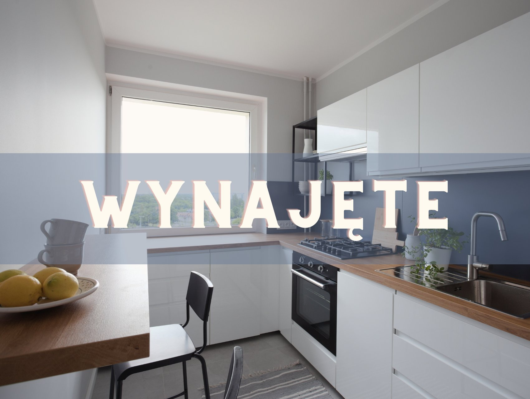 Jasna kuchnia z białymi szafkami i drewnianym blatem, stół z cytrynami i filiżankami, okno z widokiem na zieleń. Na zdjęciu widnieje napis: Wynajęte.