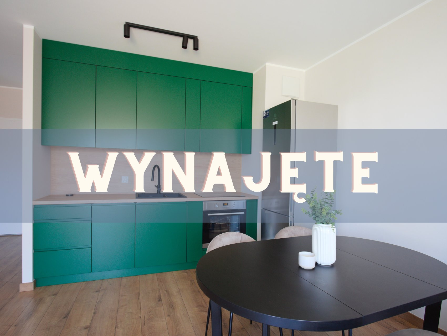 Wnętrze z zieloną zabudową kuchenną i czarnym stołem. Na blacie wazon z rośliną. Na zdjęciu widnieje napis: Wynajęte.