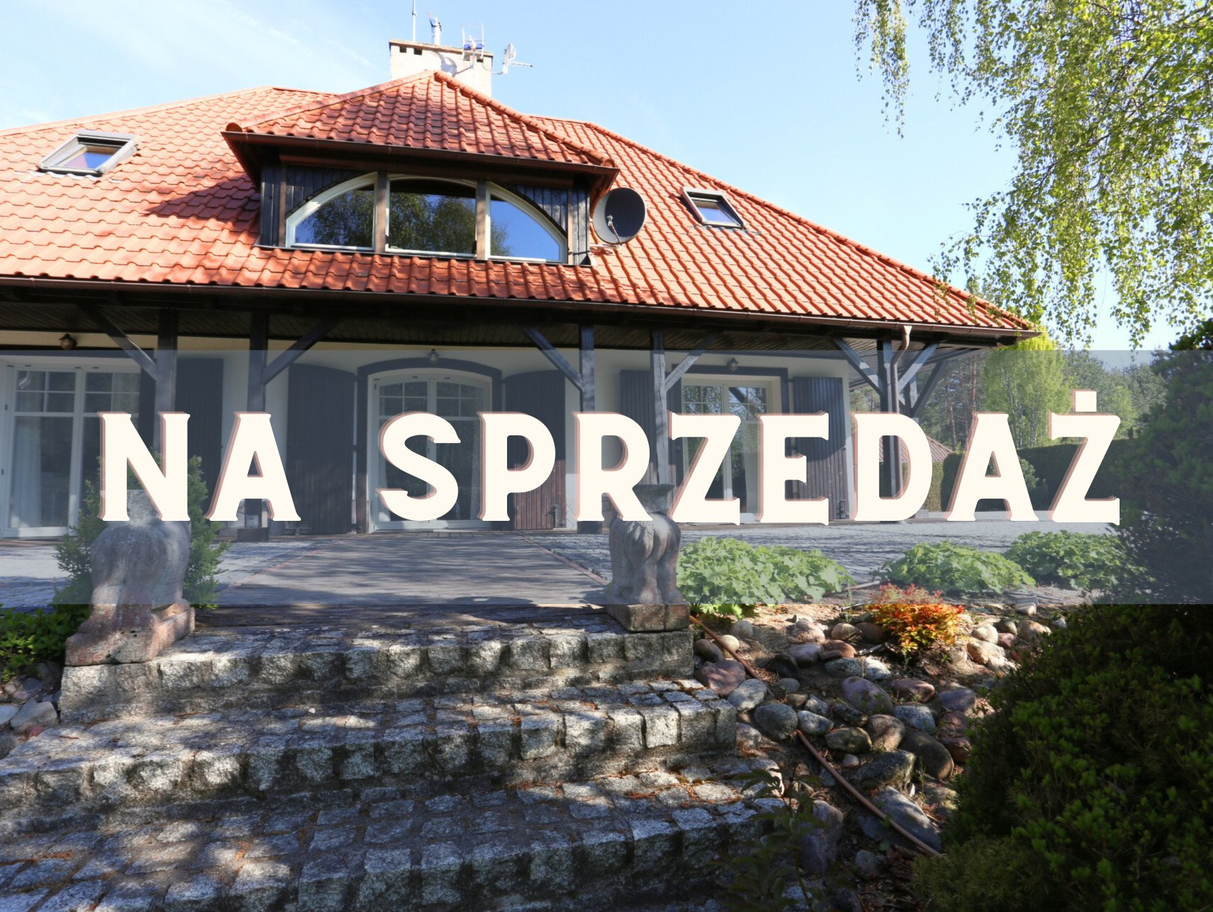 Dom z czerwonym dachem i napisem 'Na Sprzedaż' na pierwszym planie. Widoczne kamienne schody i zieleń. Słoneczny dzień, błękitne niebo.