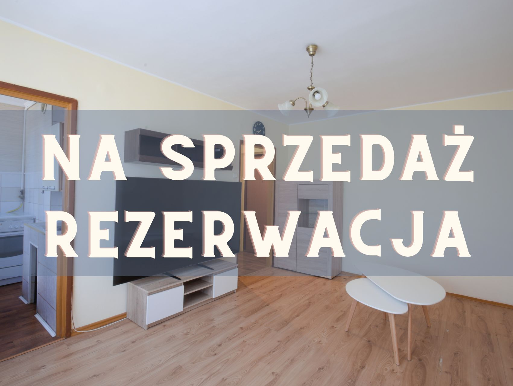 Salon z parkietem, meblami i napisem 'Na Sprzedaż Rezerwacja'. Widok na kuchnię przez otwarte drzwi. Żyrandol na suficie. Ustawiony stół kawowy.