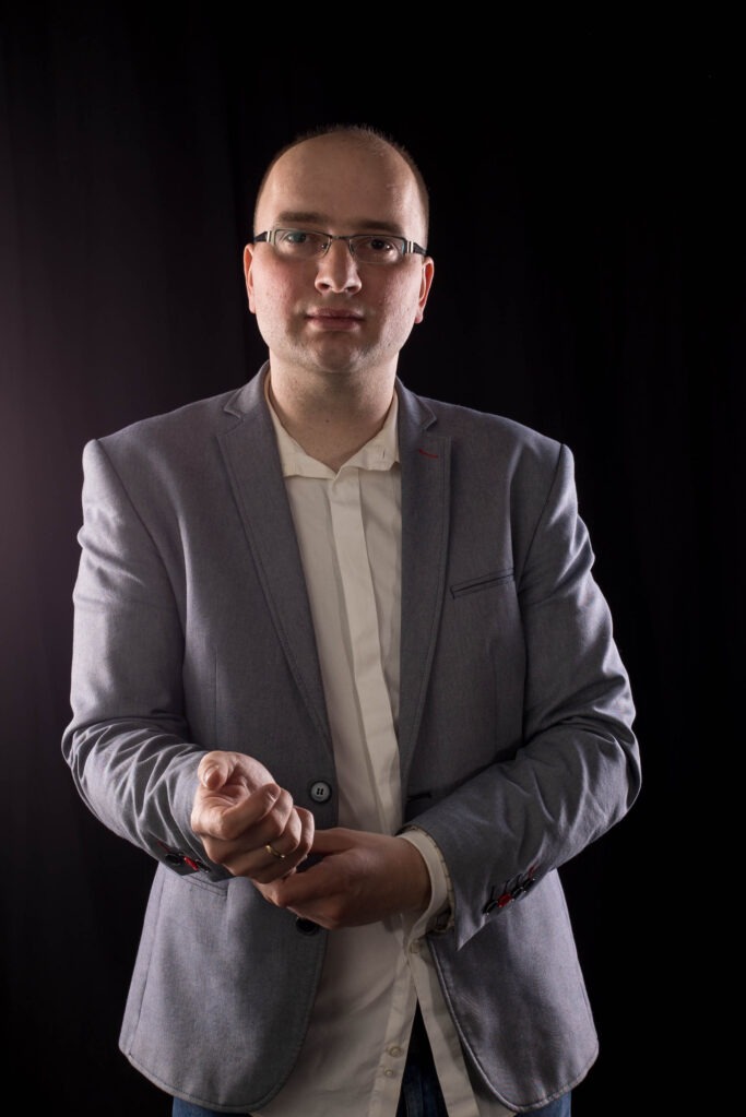 Mężczyzna w okularach i szarym garniturze na czarnym tle, delikatnie poprawia mankiet koszuli, prezentując profesjonalny wizerunek.