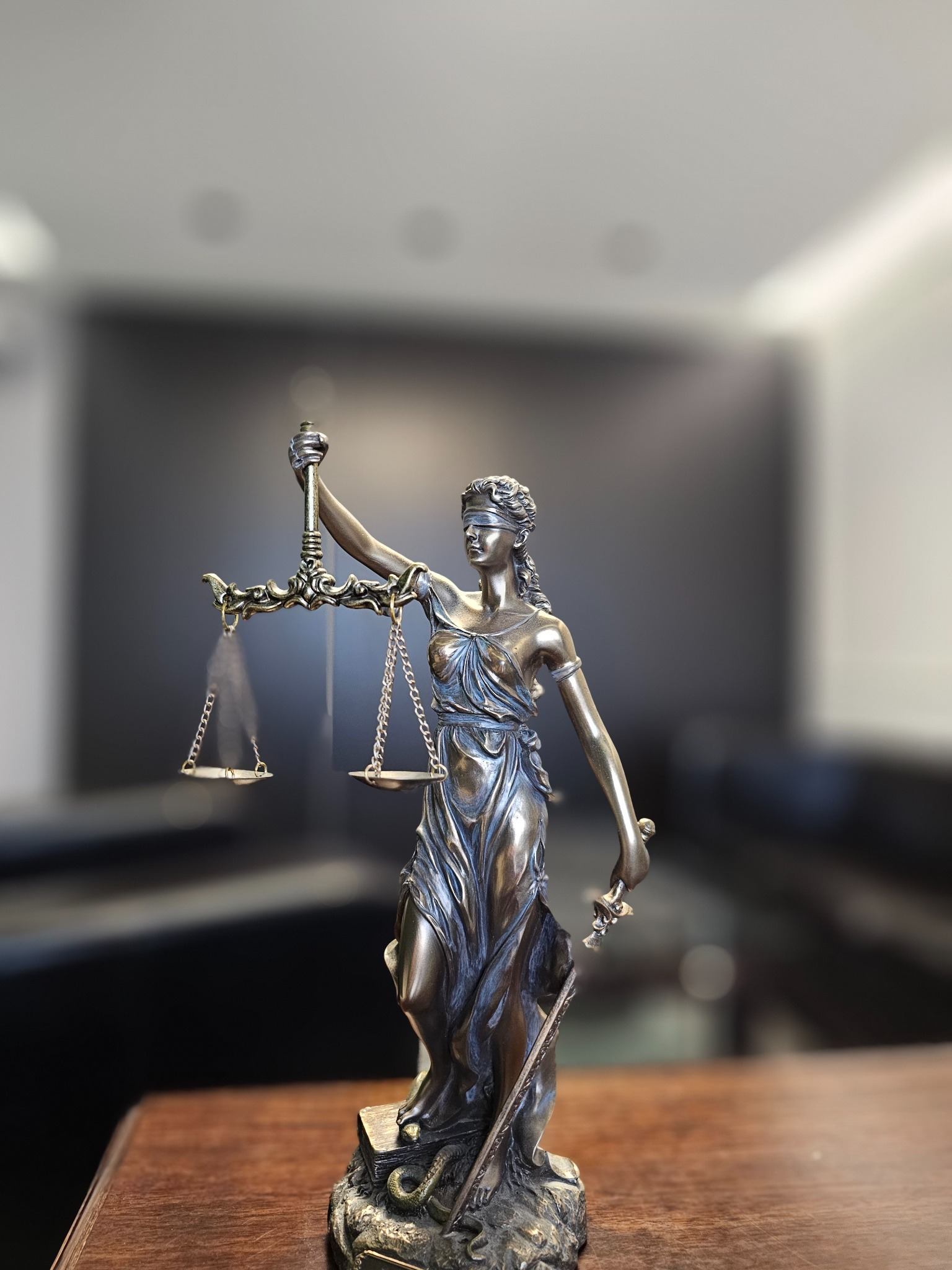 Temida, bogini sprawiedliwości, z wagą i mieczem, na drewnianym stole w kancelarii prawnej. Statuetka z brązu na tle rozmytego wnętrza biura.