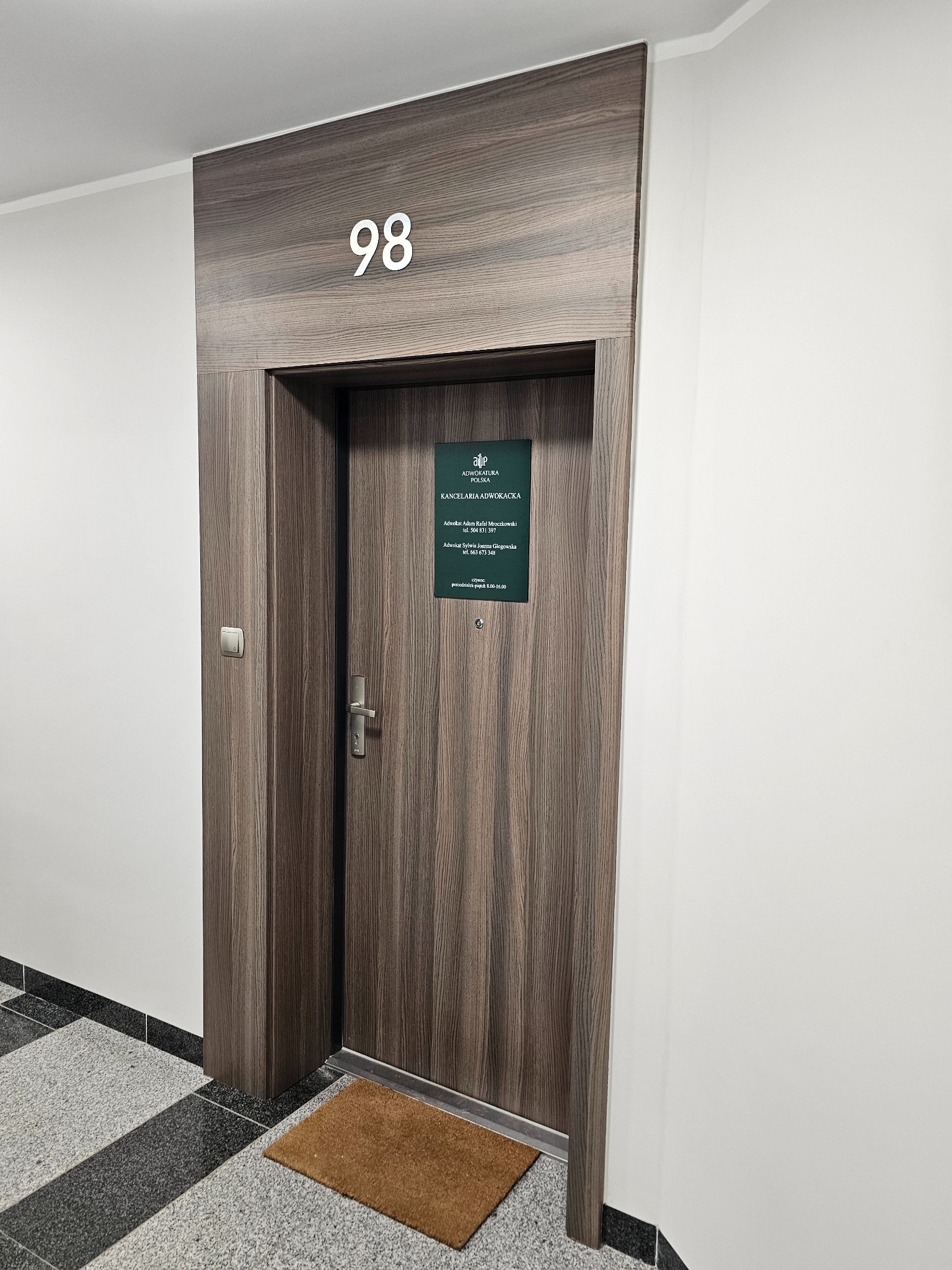 Wejście do Kancelarii Adwokackiej nr 98 z tabliczką informacyjną i wycieraczką. Drewniane drzwi z numerem lokalu na górze.
