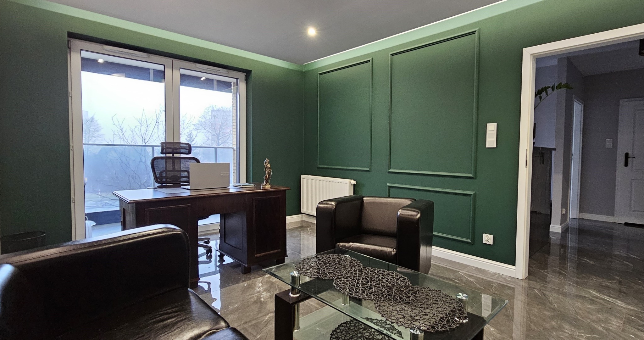 Eleganckie biuro z zielonymi ścianami, ciemnymi meblami, biurkiem z laptopem i figurką Temidy. Widok z okna na drzewa. Marmurowa podłoga i szklany stolik kawowy.