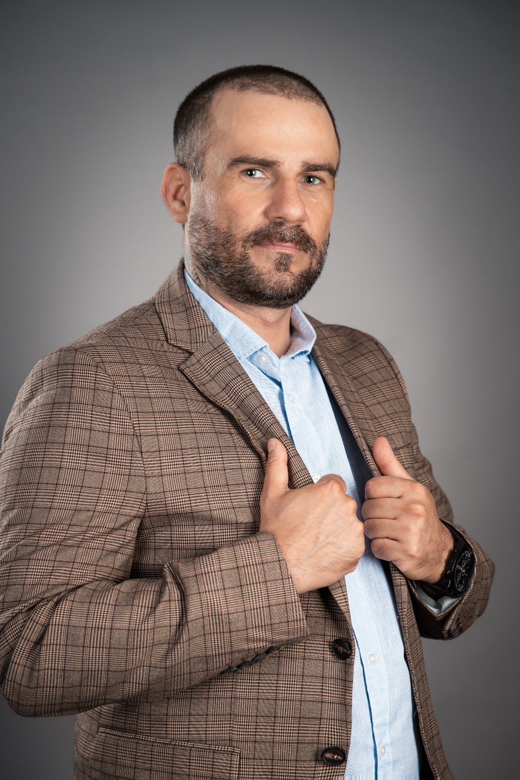 Mężczyzna z brodą w brązowej marynarce w kratę i błękitnej koszuli, pozujący na szarym tle. Widoczny zegarek na ręce.