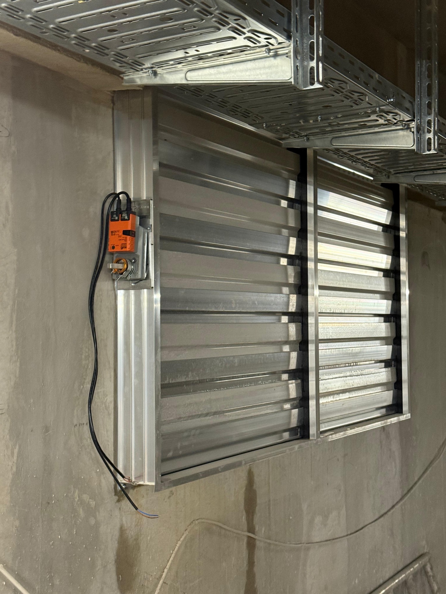 Zamontowany, metalowy system wentylacyjny z siłownikiem, widoczny na tle surowej ściany i metalowych korytek kablowych pod sufitem. Instalacja w trakcie podłączania.