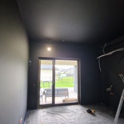 Kreatywna Przestrzeń - Pokój po malowaniu na czarno, widoczne kable i drabina. Folia ochronna na podłodze. Widok na ogród przez okno balkonowe.