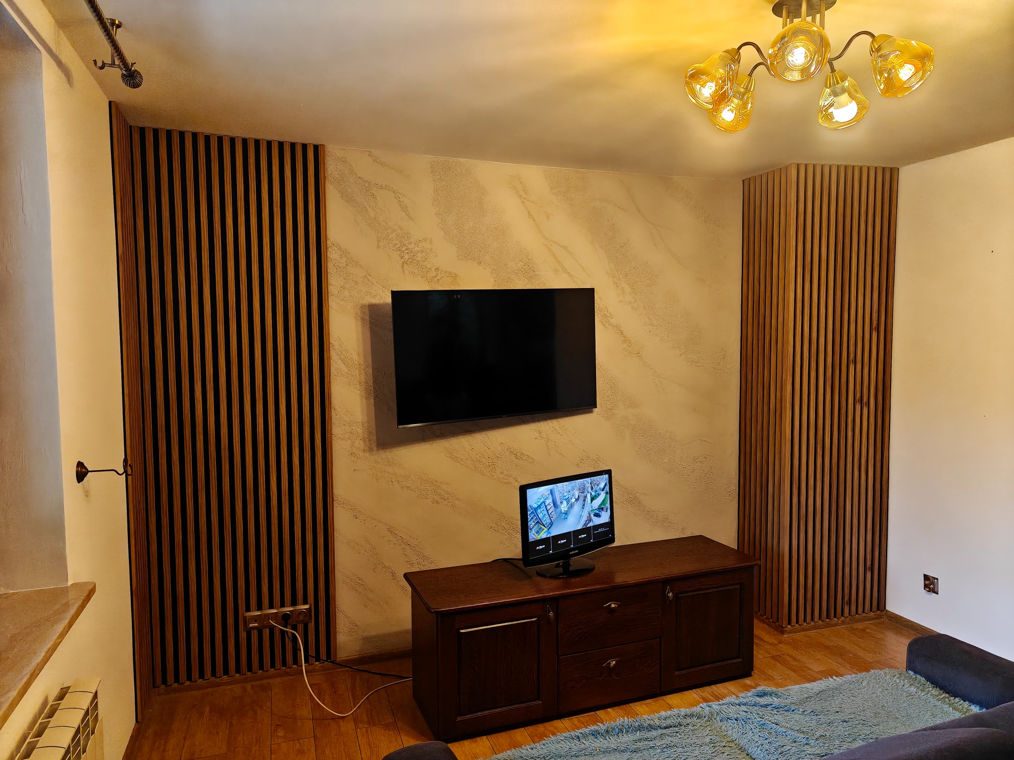 Salon po remoncie: dekoracyjne lamele ścienne z drewna, kremowa ściana z efektem dekoracyjnym, nowoczesny telewizor i drewniana komoda, ciepłe oświetlenie.