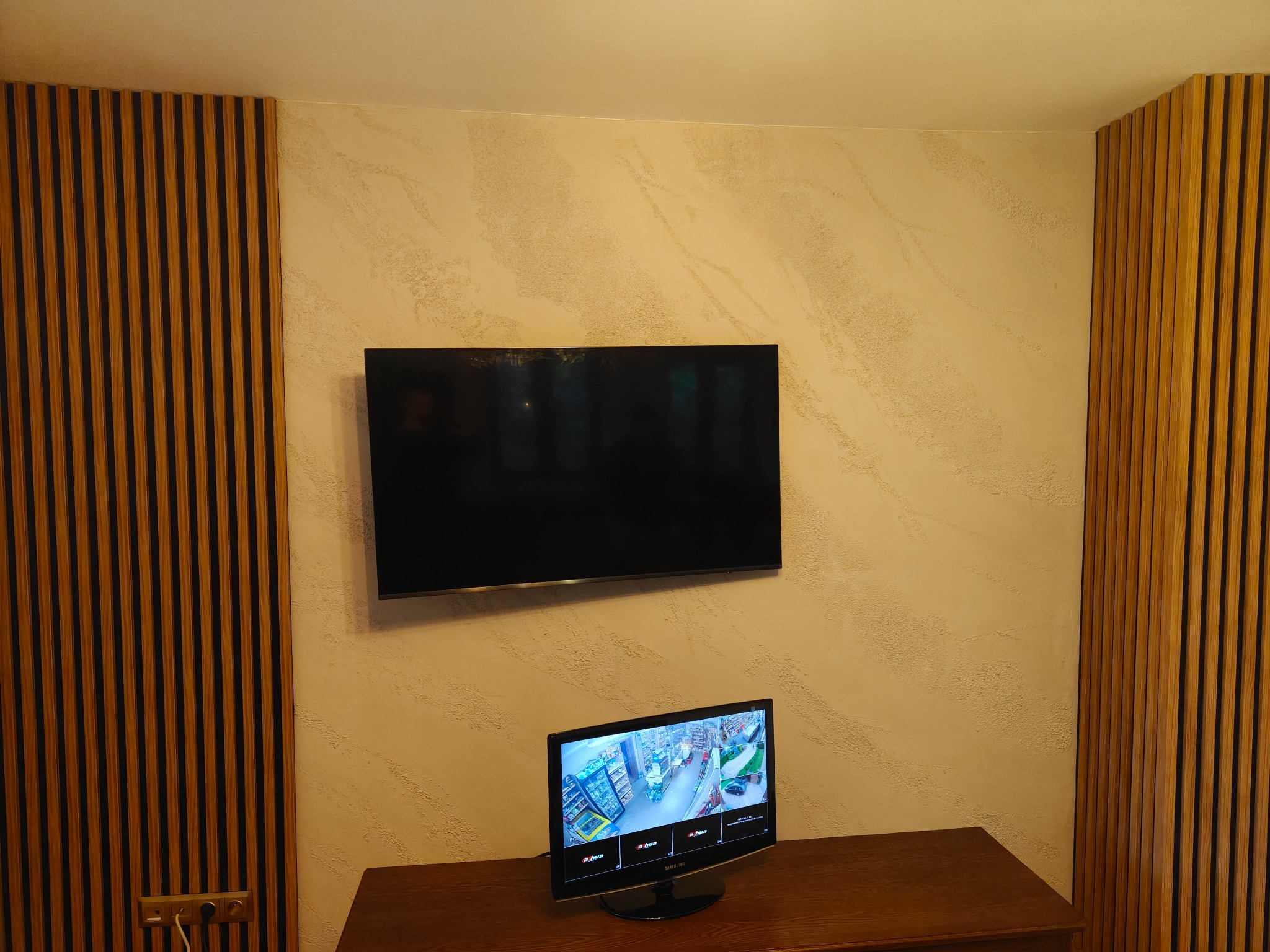 Eleganckie wykończenie ściany z dekoracyjną strukturą i drewnianymi lamelami po bokach. Na ścianie wiszący telewizor, a na komodzie mniejszy monitor.