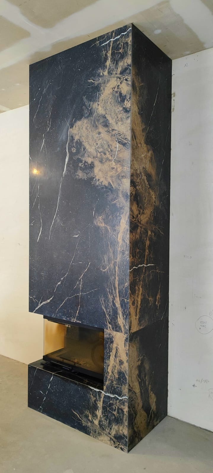 Obudowa kominka z płyt imitujących czarny marmur ze złotymi żyłami, widoczna szyba paleniska. Surowy stan wykończenia wnętrza.