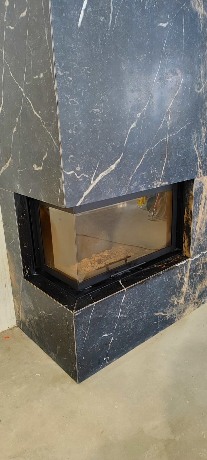 Obudowa kominka z czarnego marmuru z białymi żyłkami, widok z bliska na narożnik i szybę paleniska. Surowa podłoga w tle.