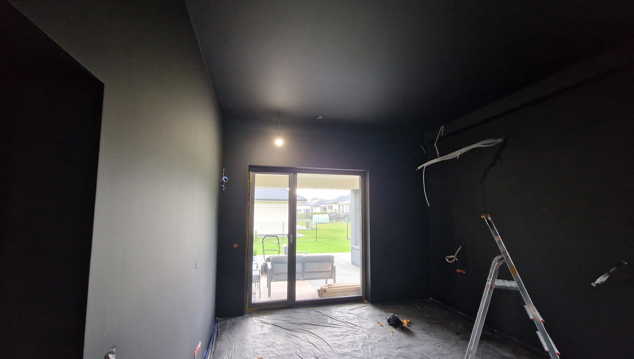 Pokój po malowaniu na czarno, widoczne kable i drabina. Folia ochronna na podłodze. Widok na ogród przez okno balkonowe.