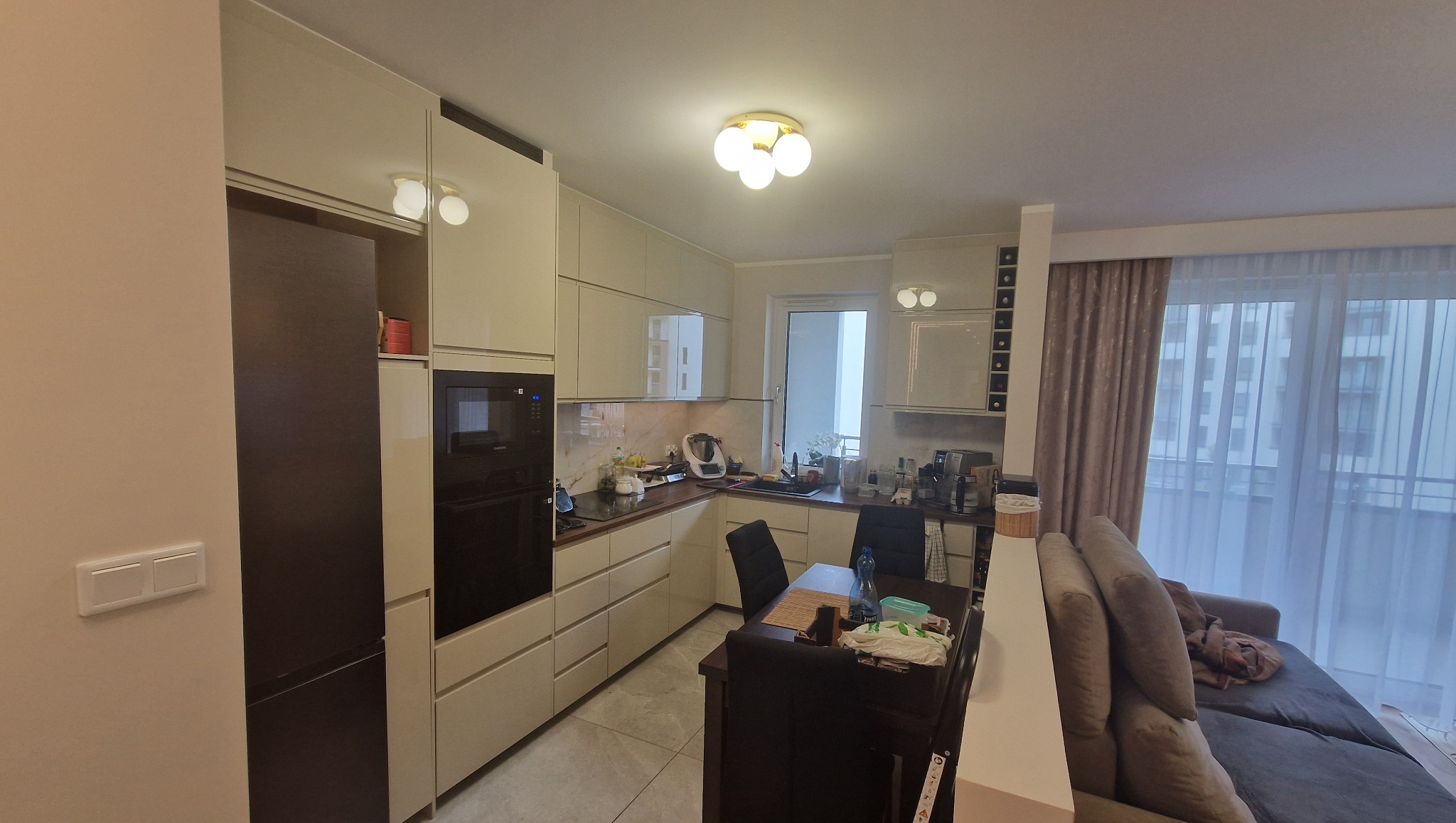 Jasna kuchnia z beżowymi szafkami i brązowym blatem, widok na salon z szarą sofą i oknem balkonowym. Nowoczesne oświetlenie sufitowe.