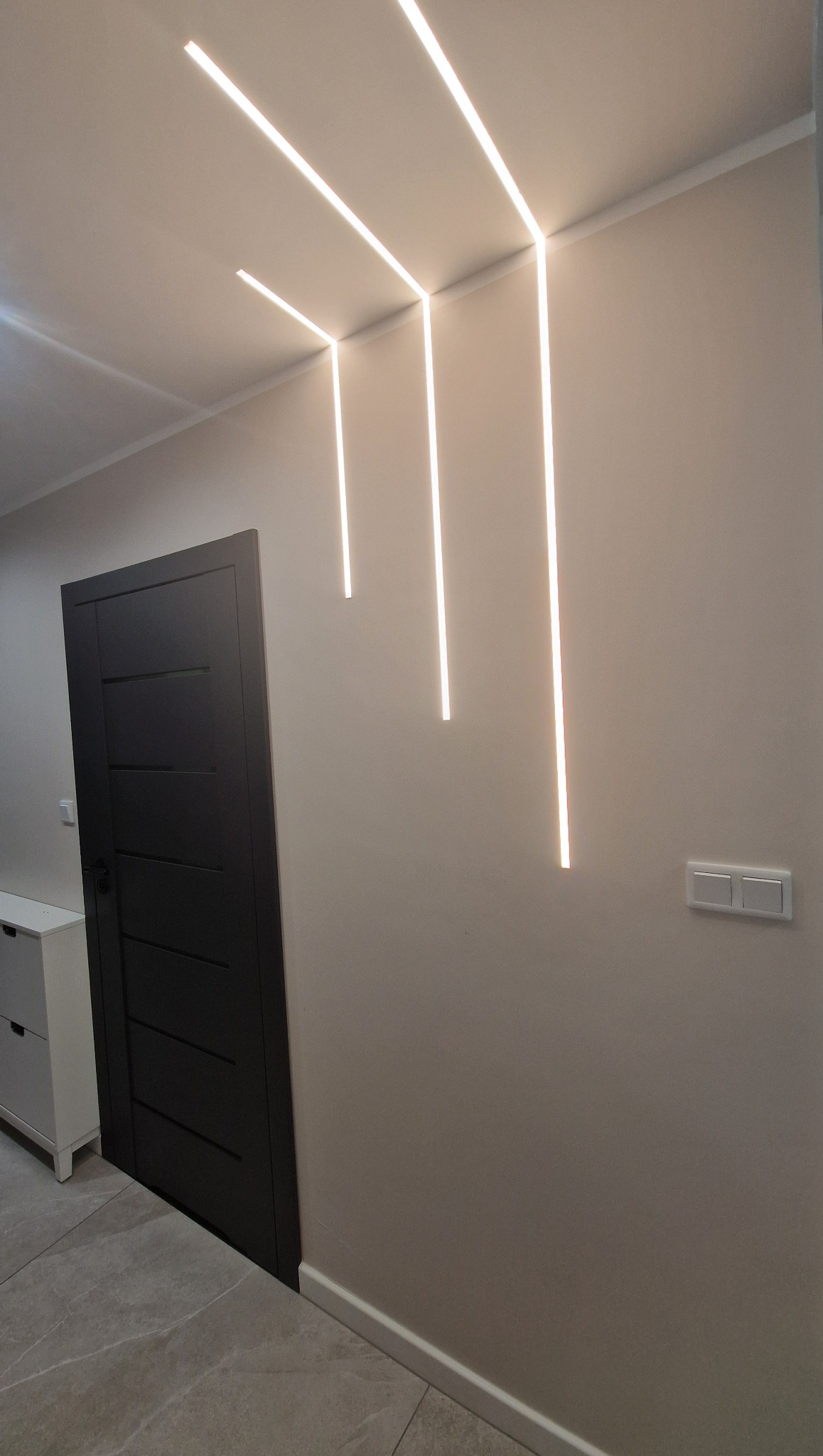 Nowoczesne oświetlenie LED wkomponowane w sufit i ścianę, tworzące geometryczny wzór. Widoczna szara ściana, drzwi i fragment komody.