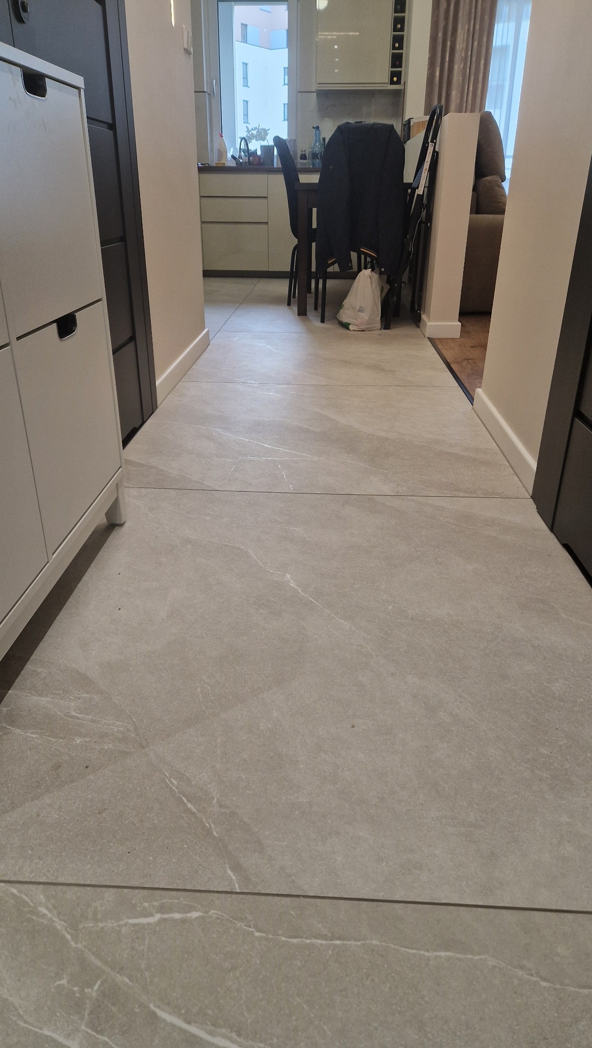 Jasnoszara podłoga z płytek imitujących marmur, ułożona w długim korytarzu łączącym kuchnię z salonem. Widoczne meble w tle i precyzyjne fugi.