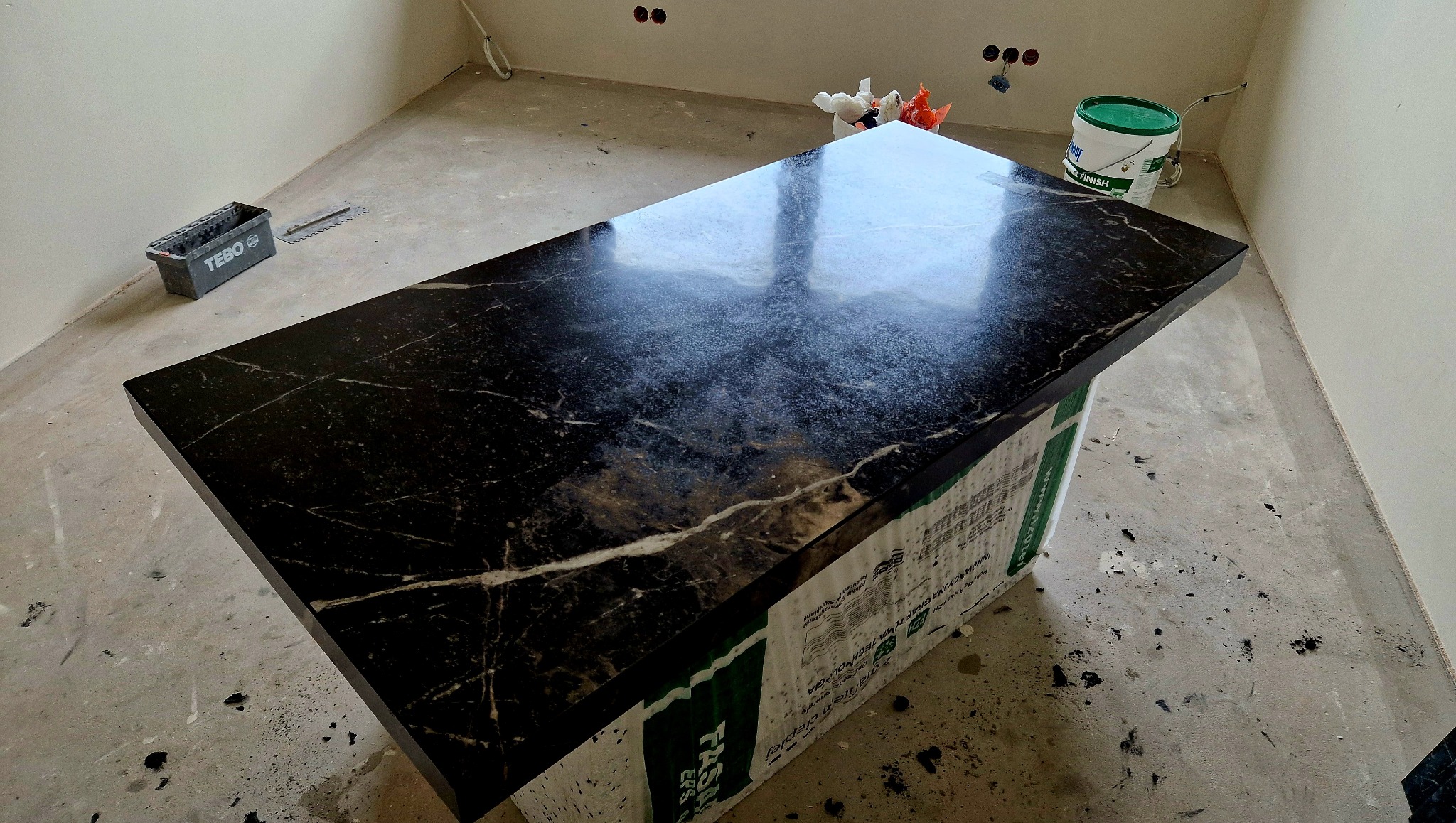 Błyszczący, czarny blat z marmurowym wzorem, oparty na zabezpieczonej konstrukcji w surowym wnętrzu. Widoczne narzędzia i wiadro farby.
