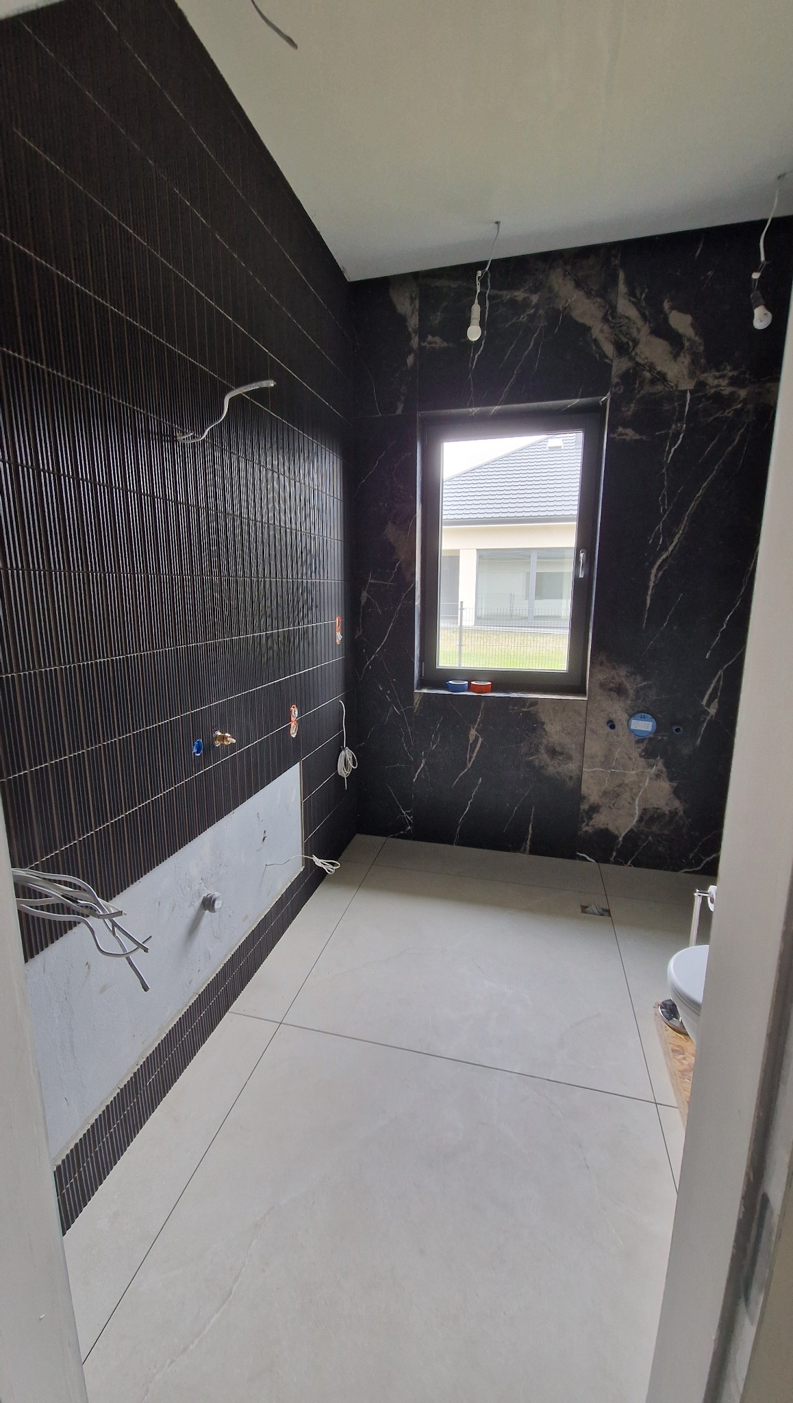 Wnętrze łazienki w trakcie prac wykończeniowych: ułożone ciemne płytki na ścianach, jasne płytki na podłodze, okno z widokiem na dom. Częściowo wykończona ściana.