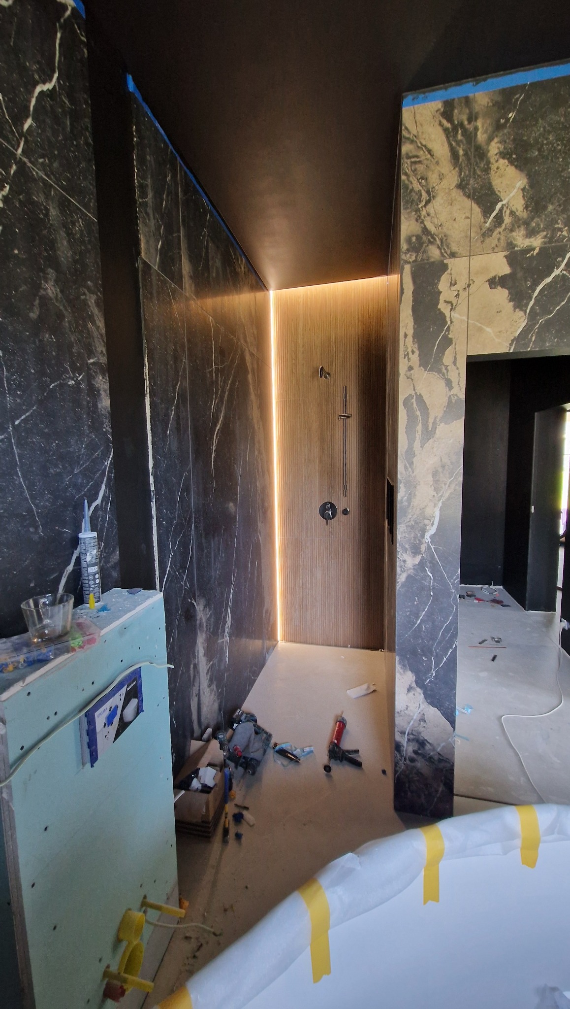 Łazienka w trakcie wykończenia: marmurowe ściany, prysznic z podświetleniem LED, widoczne narzędzia i materiały budowlane, wanna zabezpieczona folią. Prace w toku.