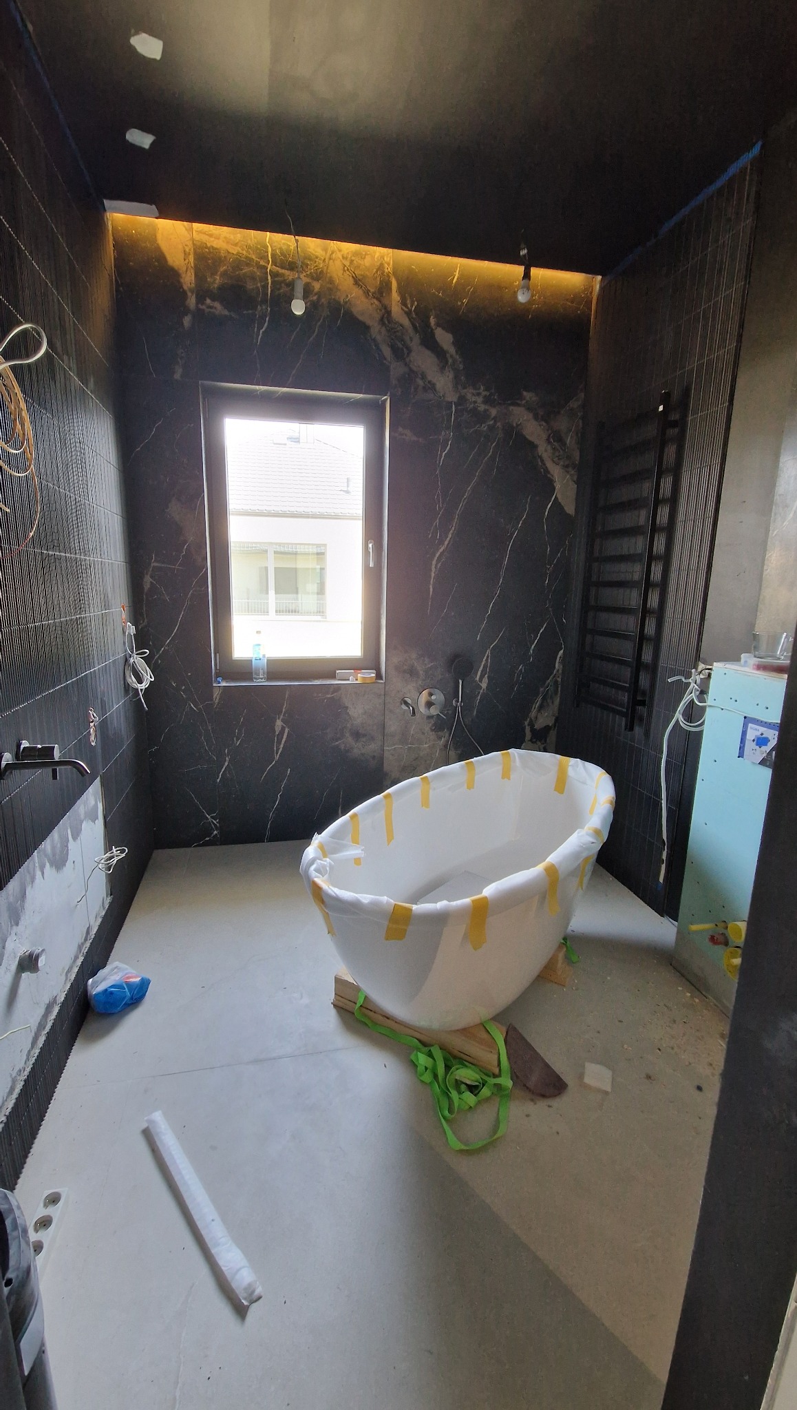 Łazienka w trakcie prac: czarne płytki imitujące marmur, okno z widokiem na dom, wanna zabezpieczona folią i taśmą, grzejnik drabinkowy.