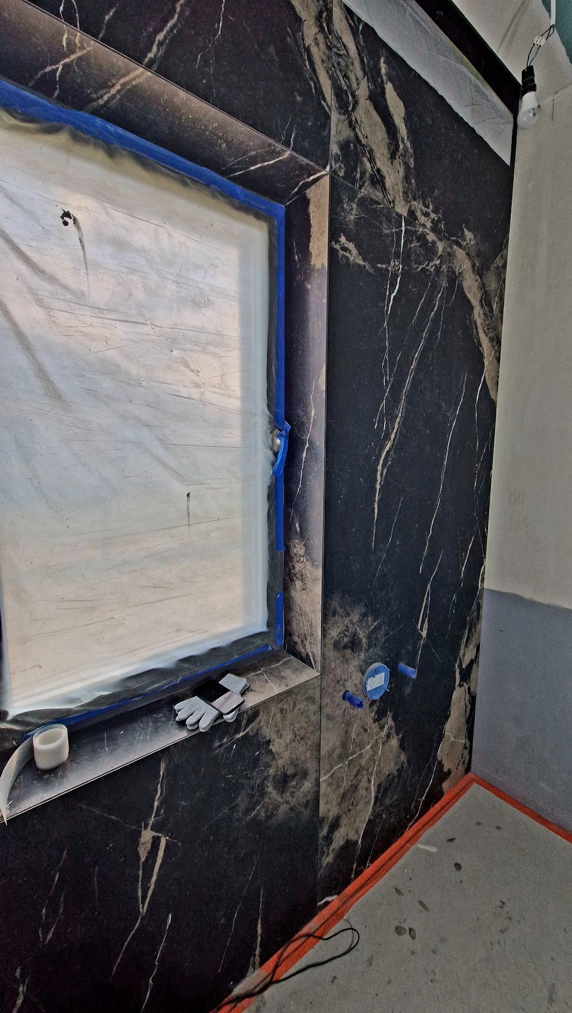 Ściana w łazience wyłożona czarnymi płytkami imitującymi marmur z widoczną ramą okna zabezpieczoną folią i taśmą malarską. Na parapecie leżą rękawice robocze.