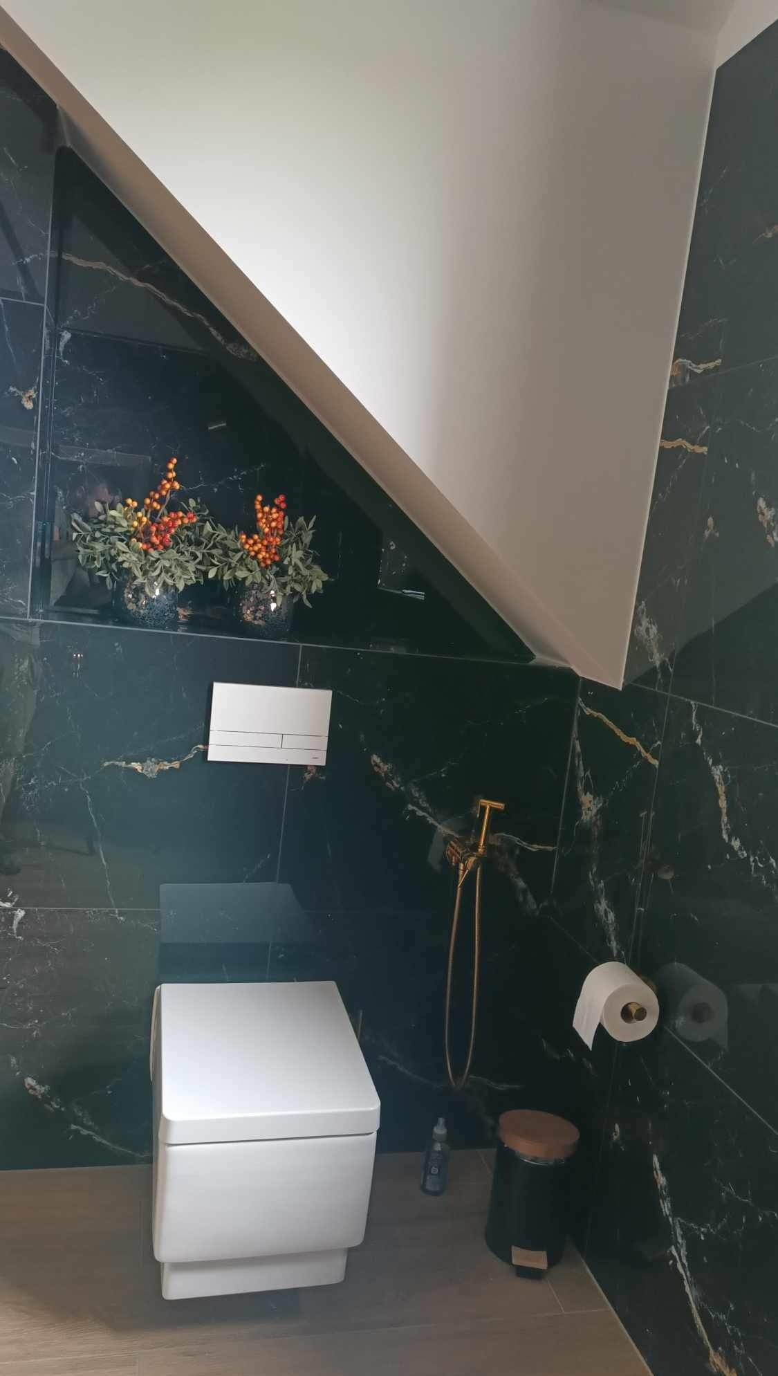 Elegancka łazienka z czarnymi płytkami imitującymi marmur, białą podwieszaną toaletą i złotą armaturą. Uwagę przyciąga skośny sufit i dekoracja z gałązek w wazonie.