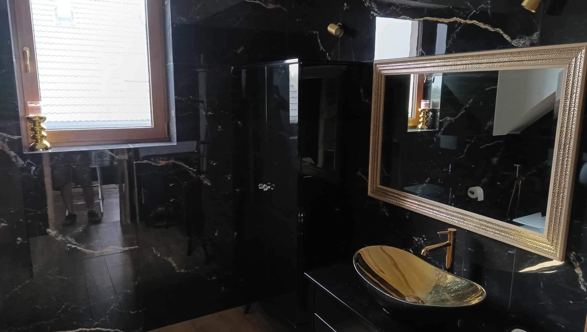 Elegancka łazienka w czerni i złocie: umywalka w kształcie łodzi, lustro w bogatej ramie i okno z dekoracyjnym świecznikiem. Luksusowe wykończenie z marmurowymi płytkami.