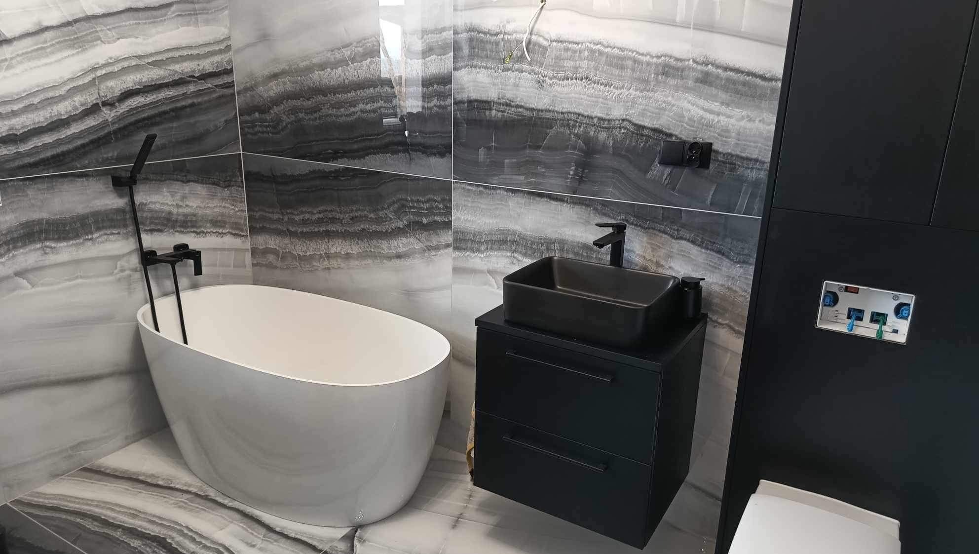 Elegancka łazienka z wanną wolnostojącą i czarną szafką z umywalką na tle marmurowych płytek w odcieniach szarości i bieli. Minimalistyczny design.