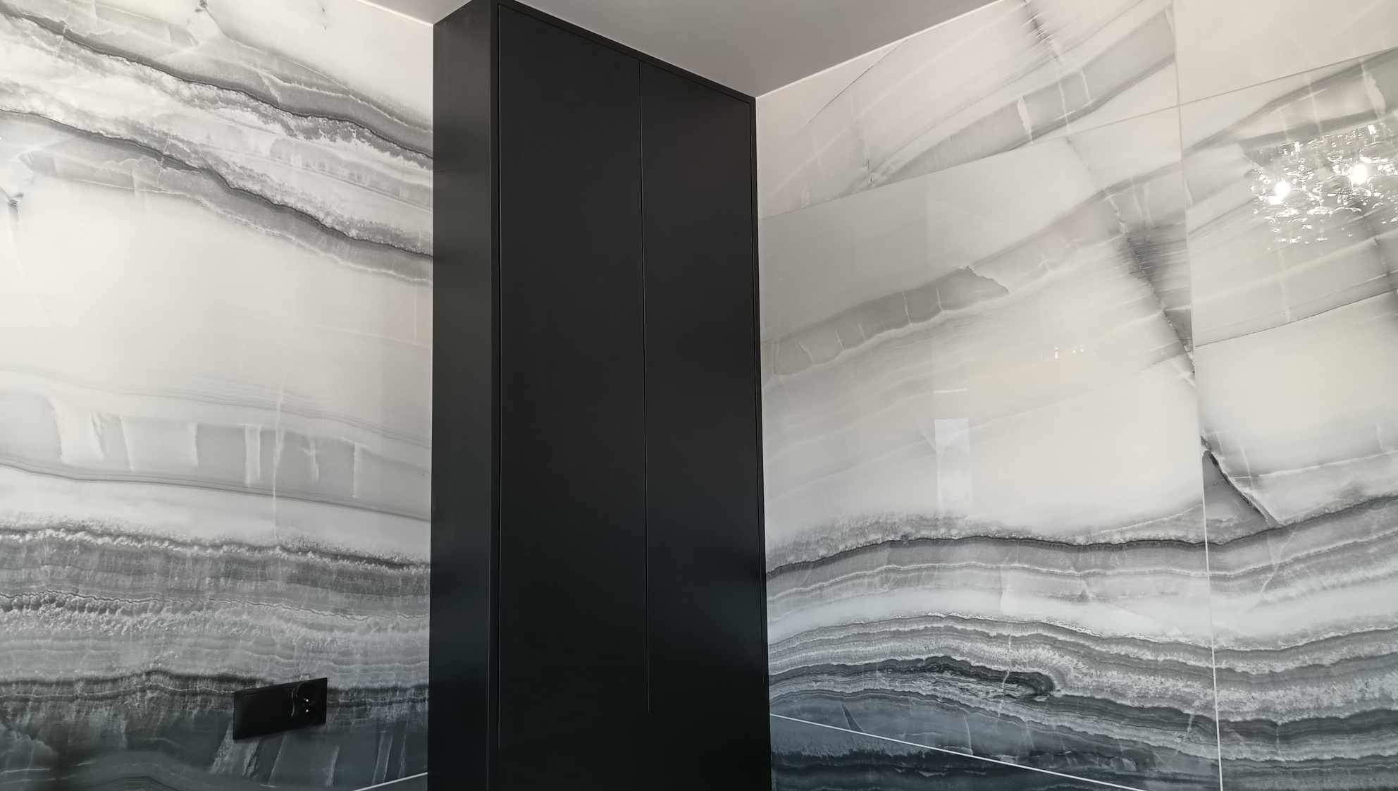 Elegancka łazienka: ściany wyłożone płytkami imitującymi onyks w odcieniach szarości i bieli, obok czarna, lakierowana szafka w zabudowie, minimalistyczny włącznik.