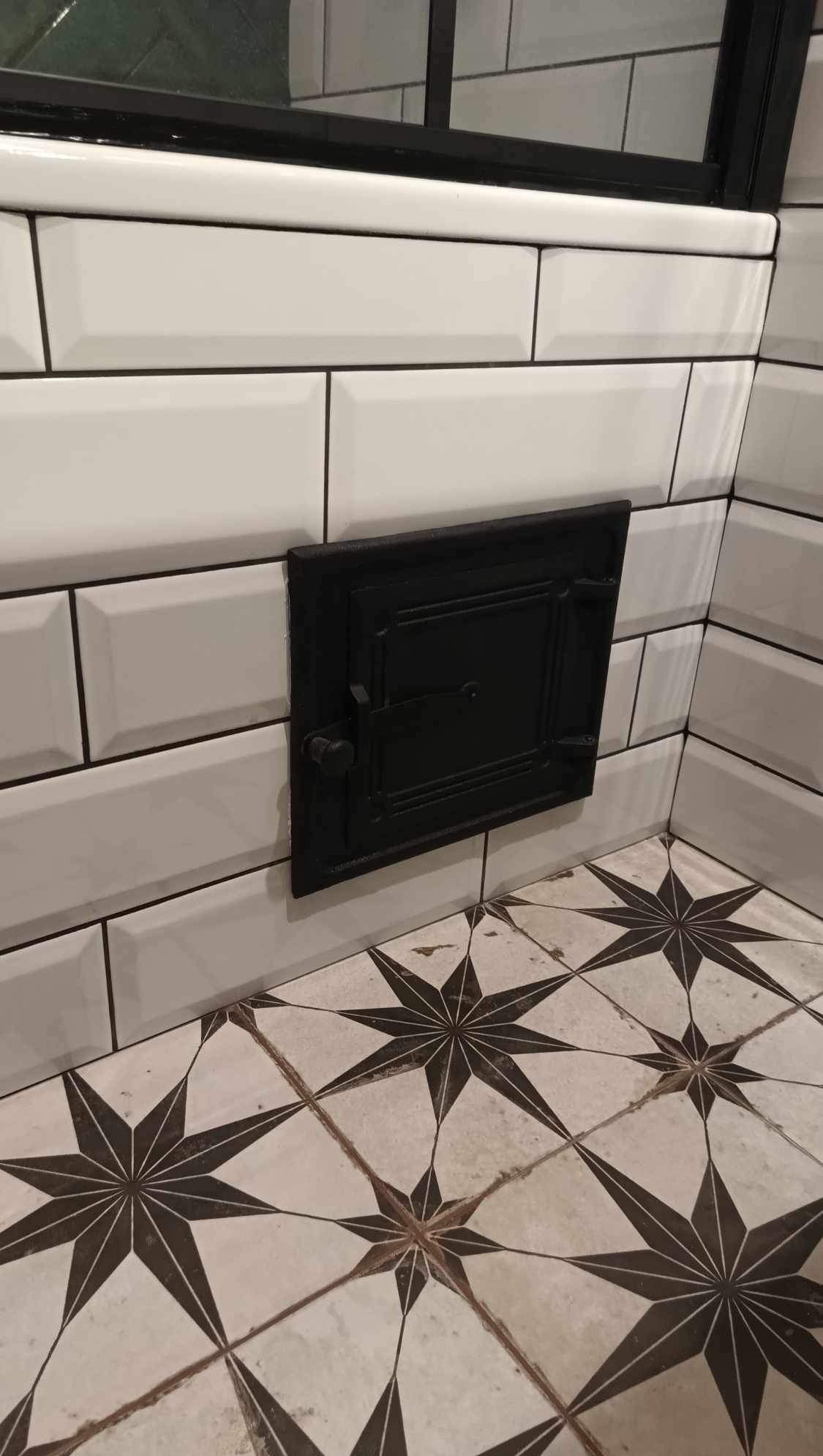 Białe, prostokątne płytki ścienne z czarną fugą, czarne drzwiczki rewizyjne i podłoga w gwiazdy. Detal wykończenia łazienki w stylu retro.