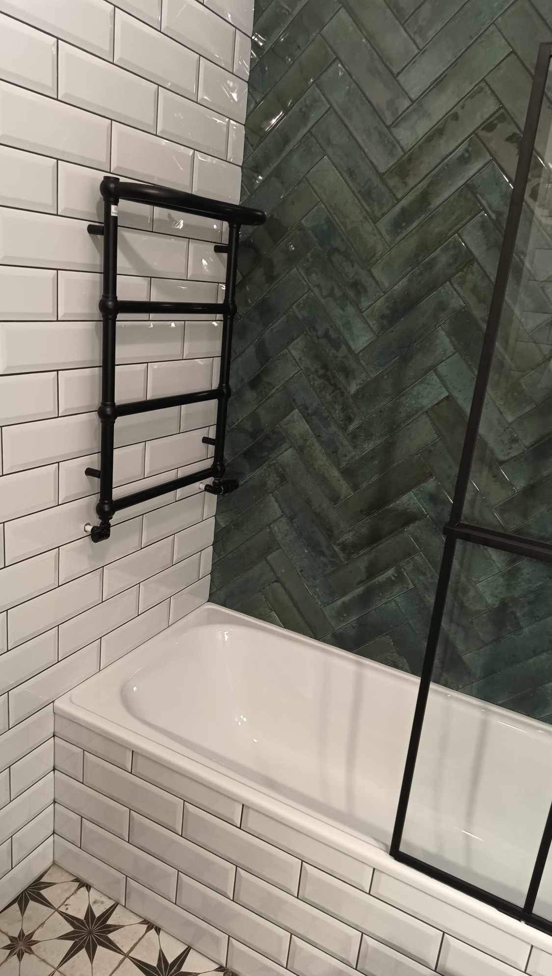 Wykończona łazienka: białe i zielone płytki ułożone w jodełkę, czarny grzejnik drabinkowy, fragment kabiny prysznicowej i wzorzysta podłoga.