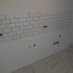 Białe, prostokątne płytki ułożone na ścianie, widoczne gniazdka i instalacja hydrauliczna. Podłoga wyłożona płytkami imitującymi marmur.