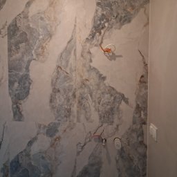 WEST-BUD IHOR MARTSYNKOVSKYI - Ściana wyłożona marmurkowymi płytkami w odcieniach szarości i beżu, z widocznymi elementami instalacji wodnej i elektrycznej. Fragment ściany pomalowany na jednolity kolor.