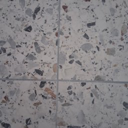 WEST-BUD IHOR MARTSYNKOVSKYI - Jasnoszara podłoga z płytek terazzo, widoczne fugi. Płytki o nieregularnym wzorze z szarymi, czarnymi i brązowymi elementami. Ujęcie z góry, wypełniające kadr.