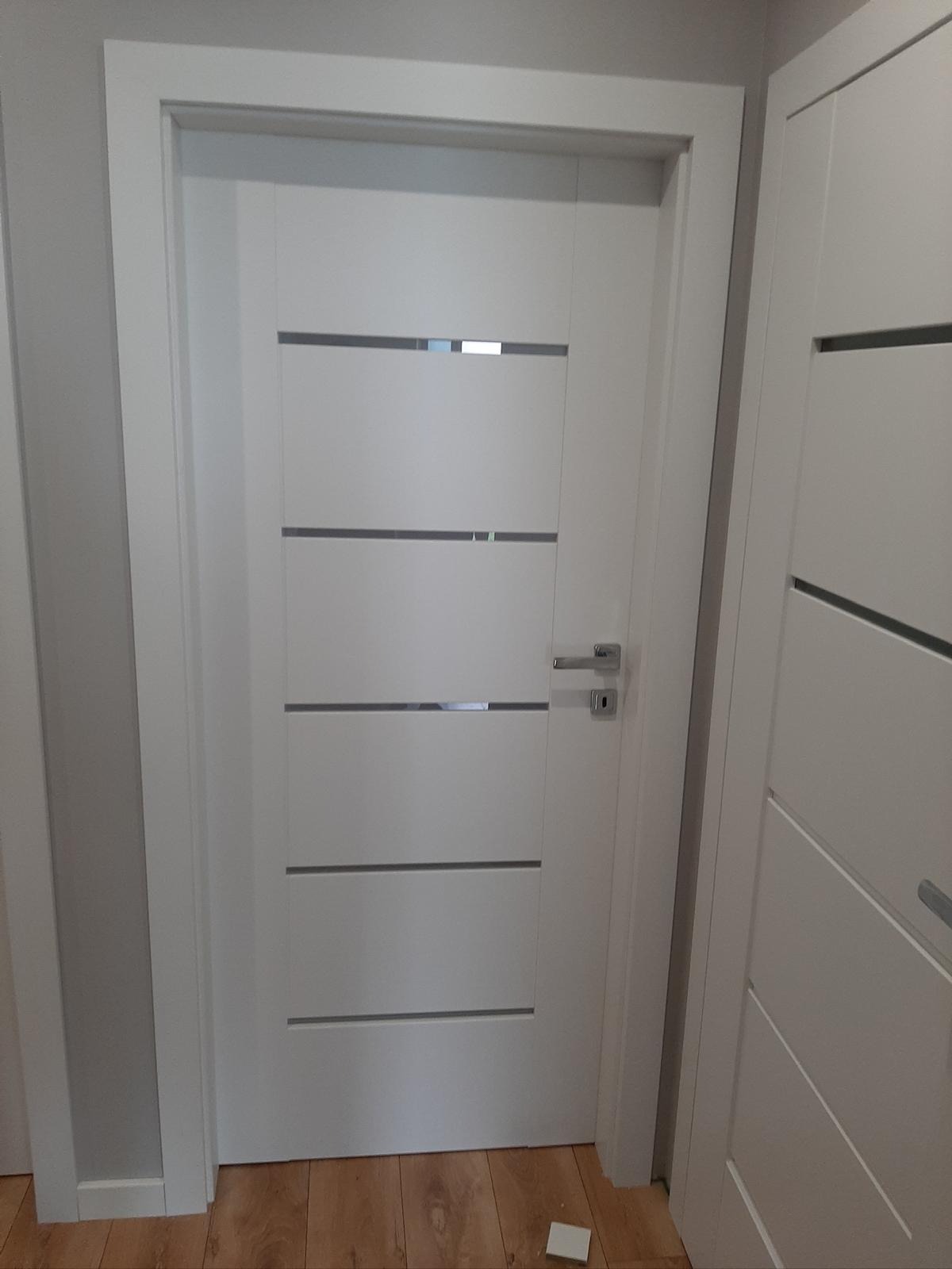 Białe, nowoczesne drzwi wewnętrzne z czterema poziomymi, szarymi wstawkami, zamontowane w ościeżnicy. Widoczny fragment drewnianej podłogi i fragment drugich drzwi.