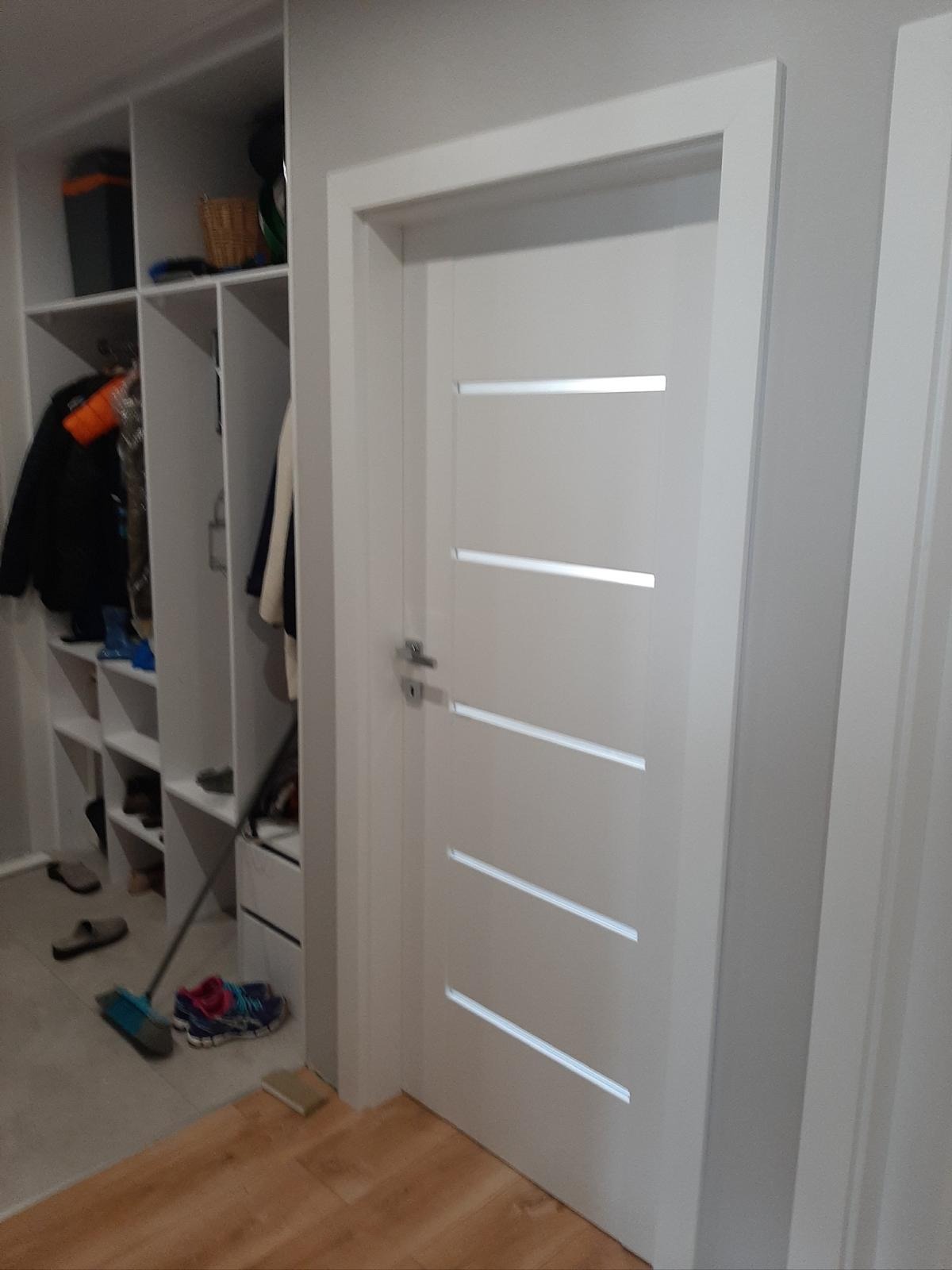 Białe drzwi wewnętrzne z poziomymi przeszkleniami, zamontowane w wejściu do garderoby z półkami i wieszakami. Podłoga drewniana, częściowo przykryta płytkami.