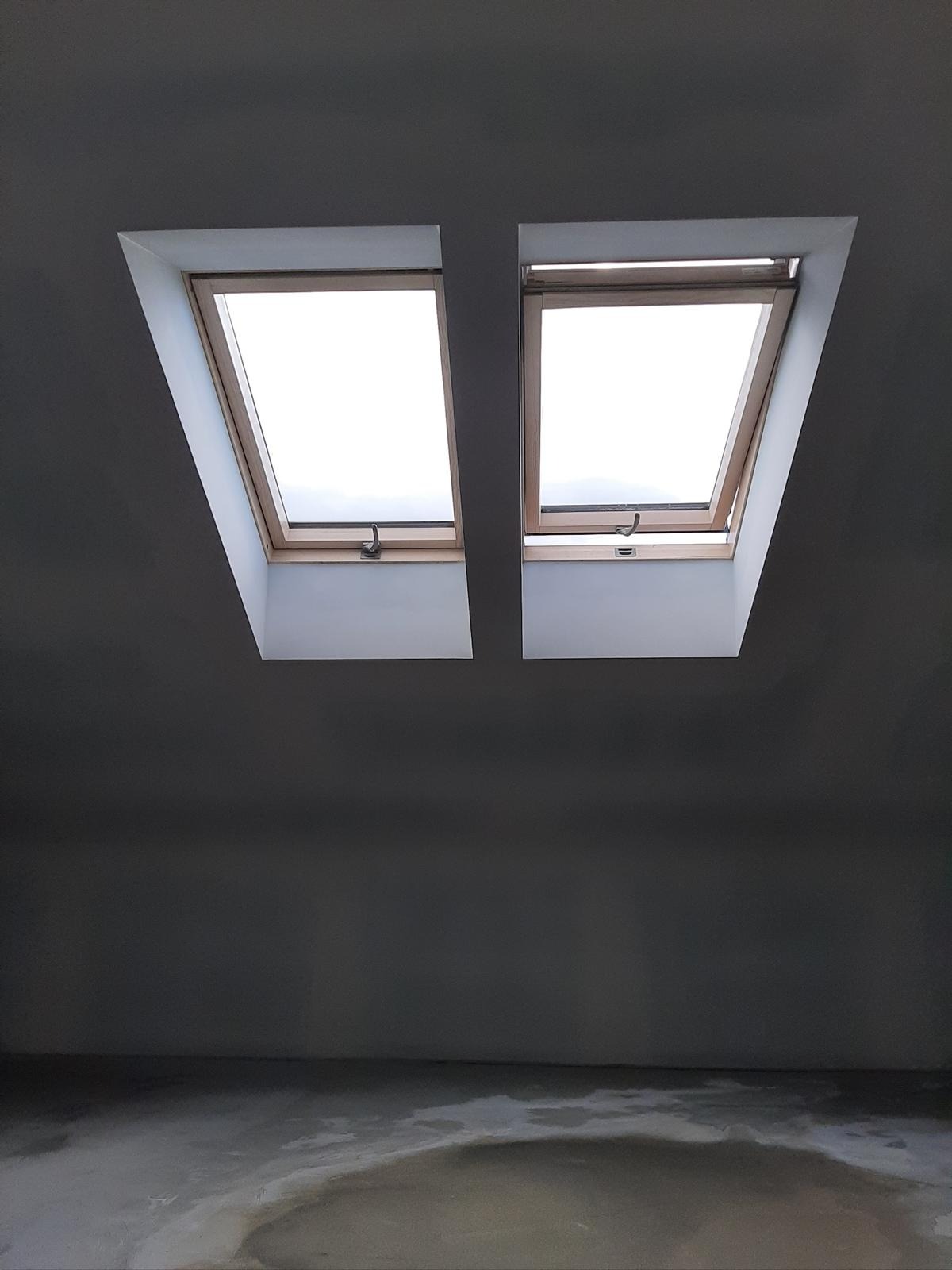 Dwa okna dachowe z drewnianą ramą w szarym pomieszczeniu na poddaszu w Warszawie. Jeden z okien jest otwarty.
