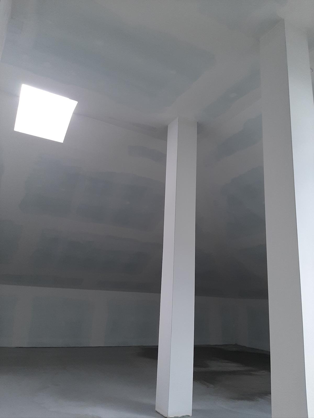 Szpachlowana sufit i ściany poddasza z oknem dachowym i kolumnami, przygotowane do malowania. Widok z dołu, surowy stan wykończenia.