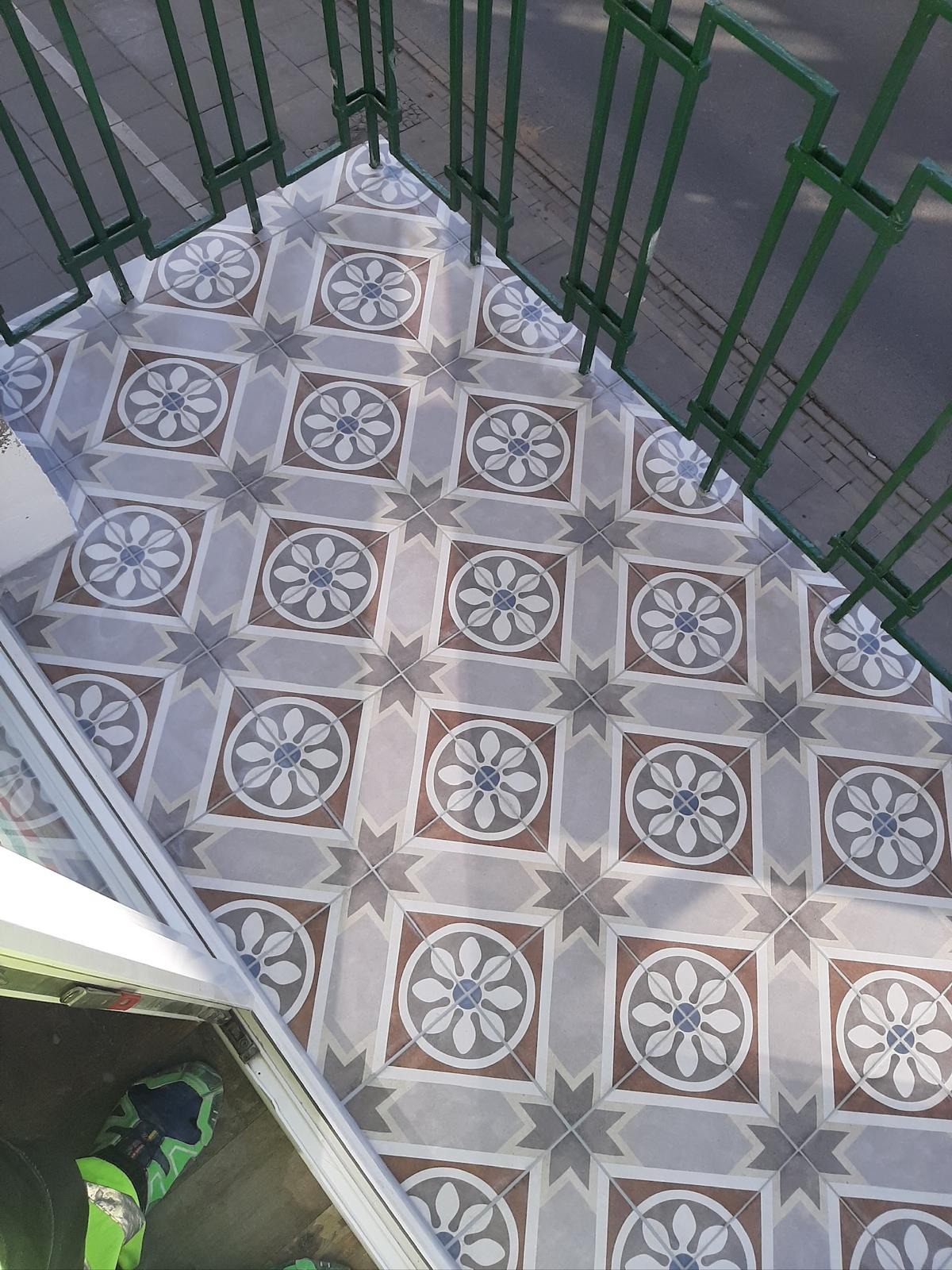 Wzorzyste płytki na balkonie z zieloną barierką, widok z góry. Geometryczny motyw kwiatowy w odcieniach szarości, brązu i bieli. Częściowo widoczny but w dolnym rogu.