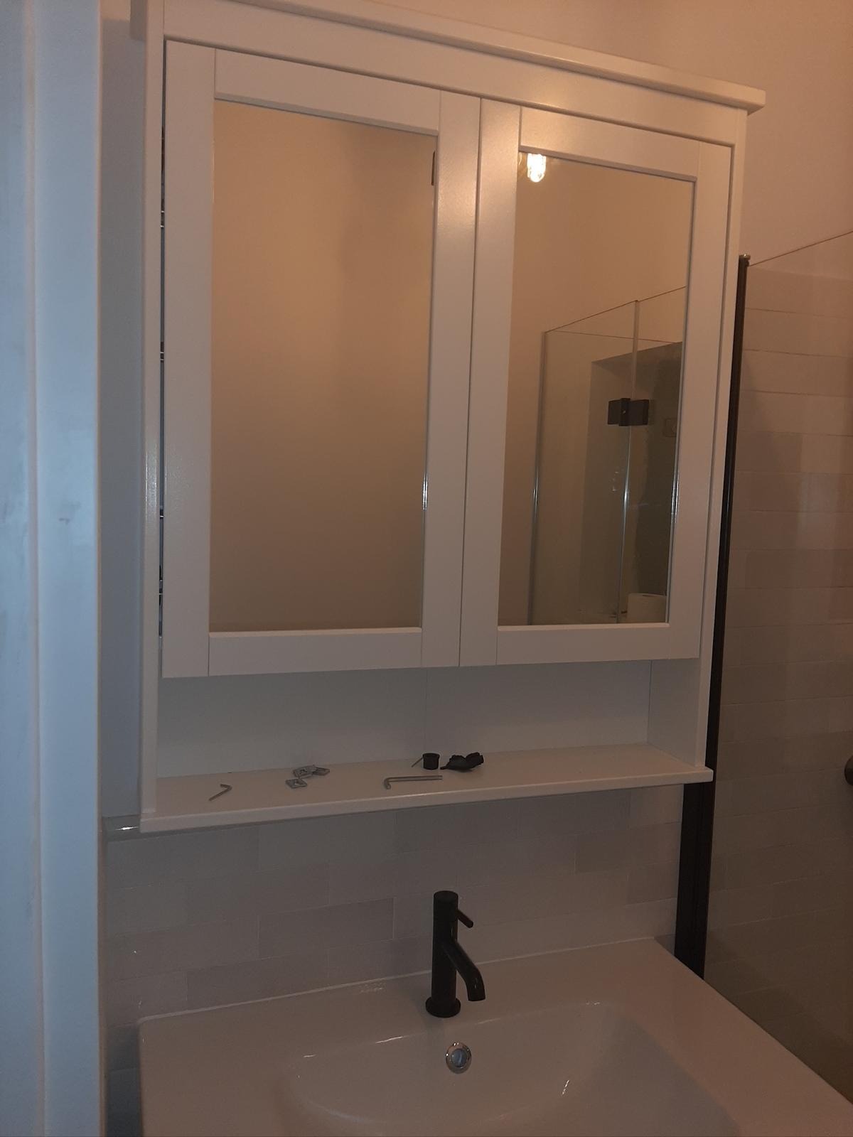 Biała szafka z lustrem nad umywalką w łazience, widoczne narzędzia na półce, czarna bateria umywalkowa. Świeżo po montażu, widok na kabinę prysznicową w odbiciu.