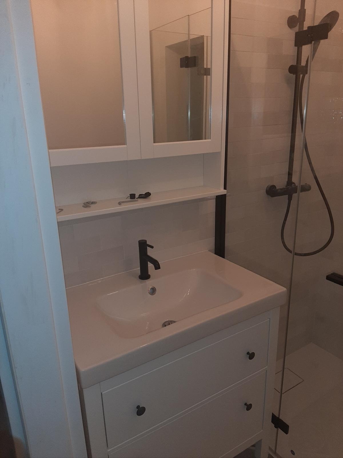 Wykończona łazienka: biała szafka pod umywalkę z czarną armaturą, lustro w szafce nad umywalką, kabina prysznicowa z ciemną deszczownicą. Minimalistyczny design.