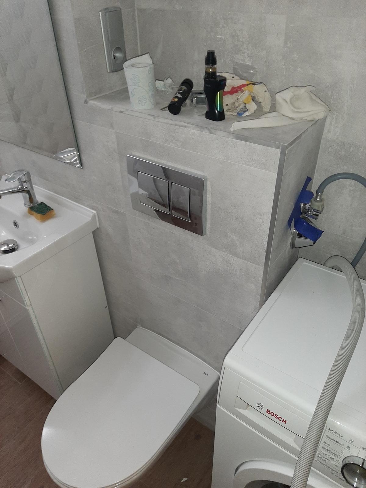 Wykończona łazienka z umywalką, toaletą i pralką Bosch. Szare płytki na ścianach, chromowane elementy. Na półce papier toaletowy i akcesoria.