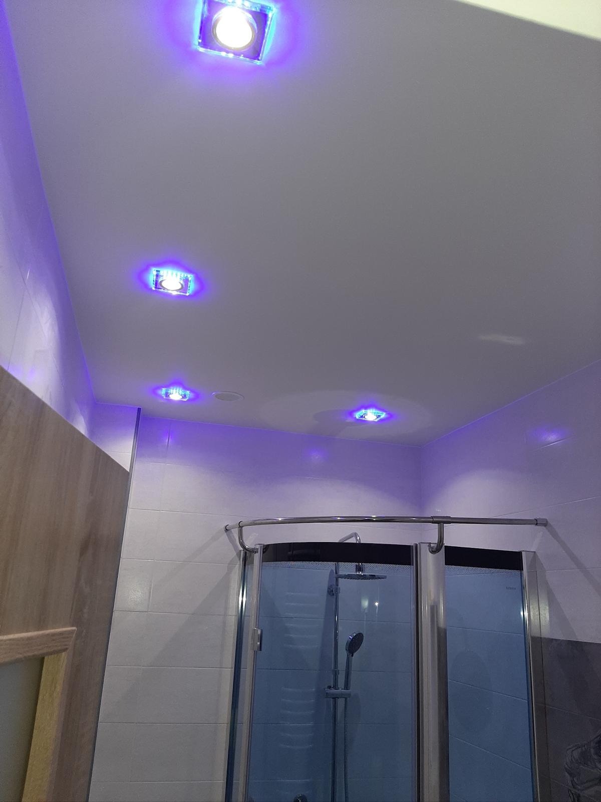 Wykończona łazienka z kabiną prysznicową, białe płytki na ścianach i suficie. Sufit podwieszany z czterema kwadratowymi lampami LED z niebieską poświatą.